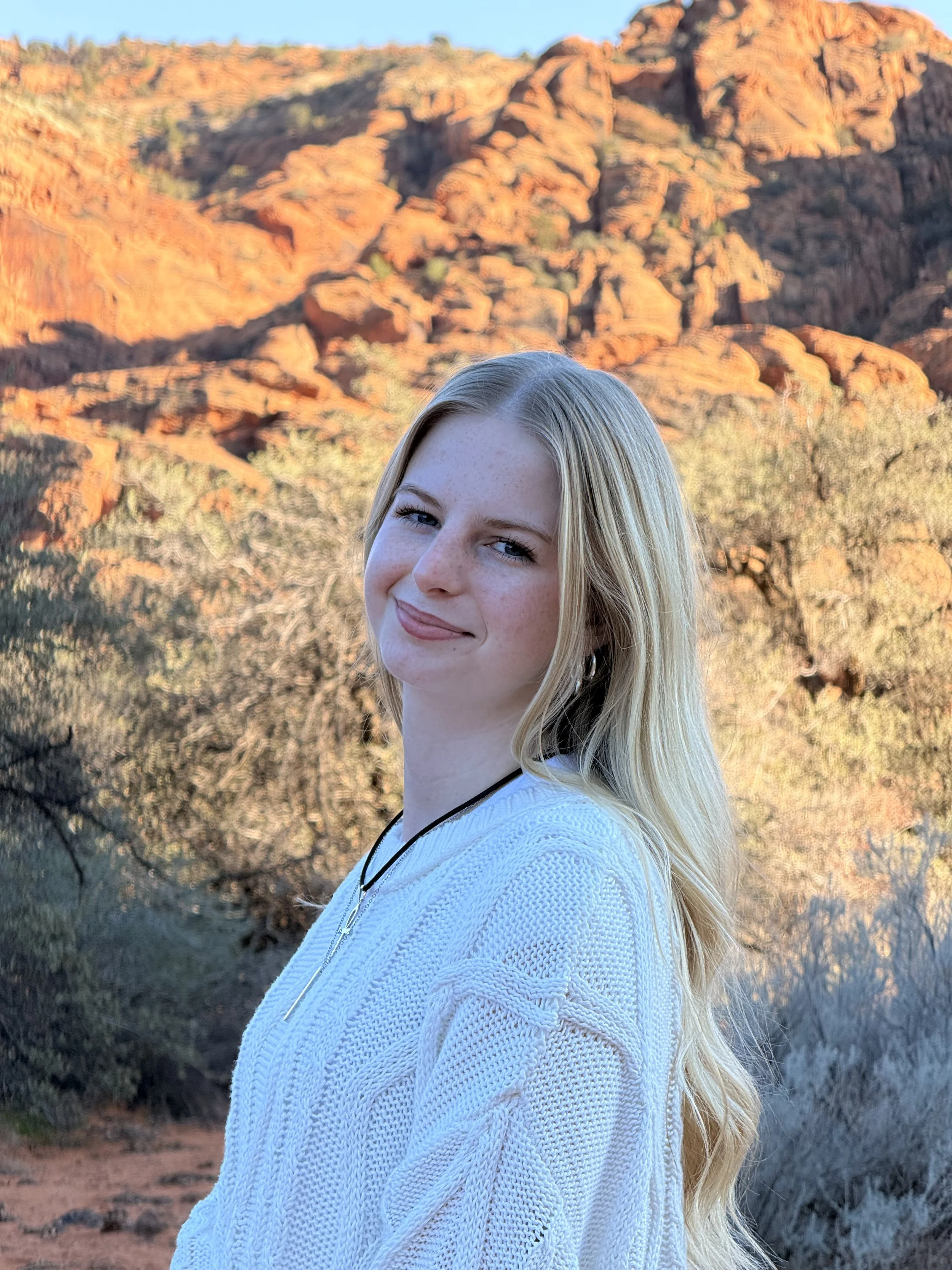 A young woman with long blonde hair and a white textured sweater smiling outdoors against a backdrop of red rocky mountains and desert vegetation.