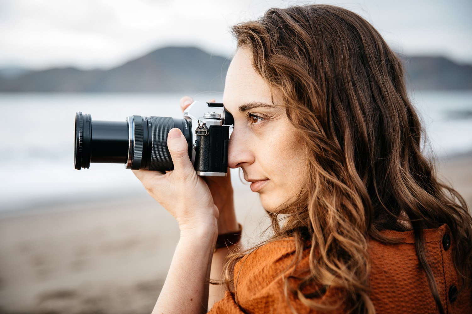  brand portrait of photographer Toni Toscano holding her Olympus camera with the ocean behind her, shot at Baker Beach in San Francisco by brand photographer Allison Busch Photography 