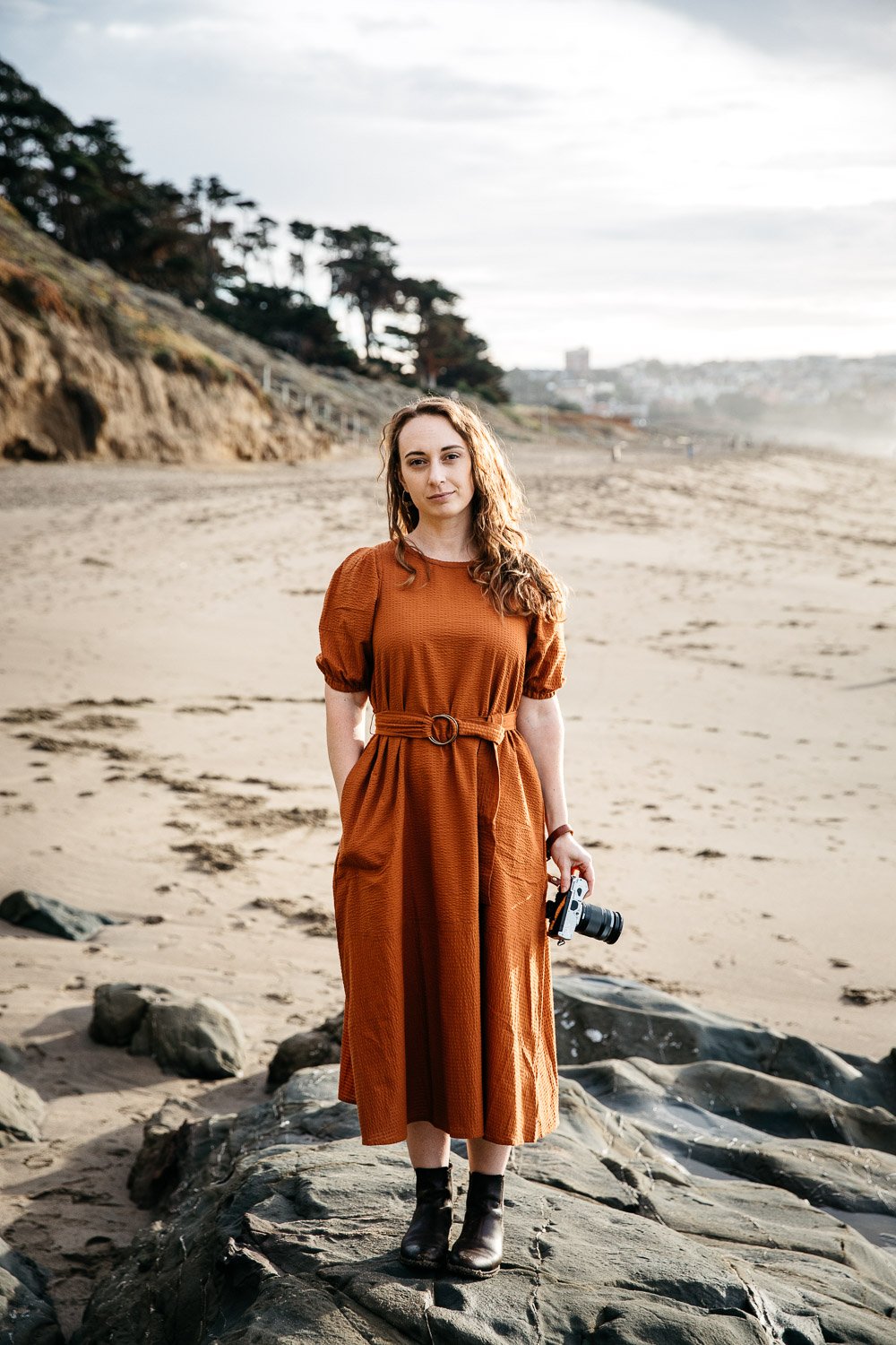  environmental portrait of photographer Toni Toscano holding her Olympus camera with a tree-lined cliff and sky behind her, shot at Baker Beach in San Francisco by brand photographer Allison Busch Photography 