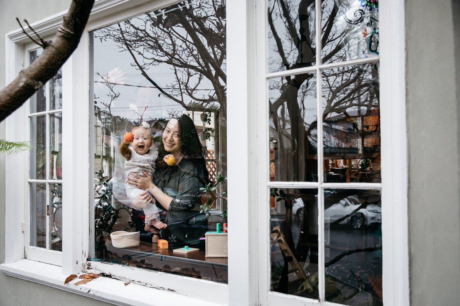  family photograph of mom, dad, and baby looking out the front window of their home, baby holds yellow and orange balls and is laughing, shot in Berkeley by family photographer Allison Busch Photography 