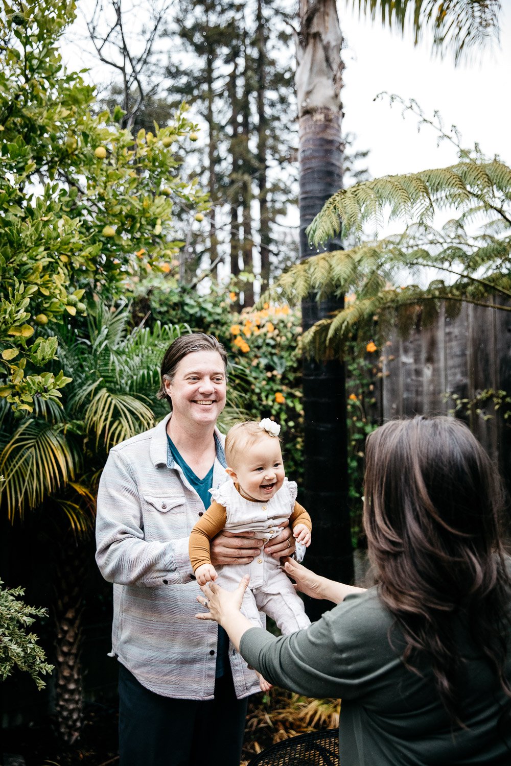  family photograph of mom, dad, and baby laughing at home in backyard filled with plants, shot in Berkeley by family photographer Allison Busch Photography 