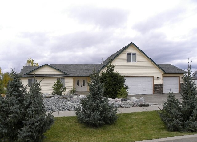  Tan home with a river rock bed, grass, and deciduous trees in front. 