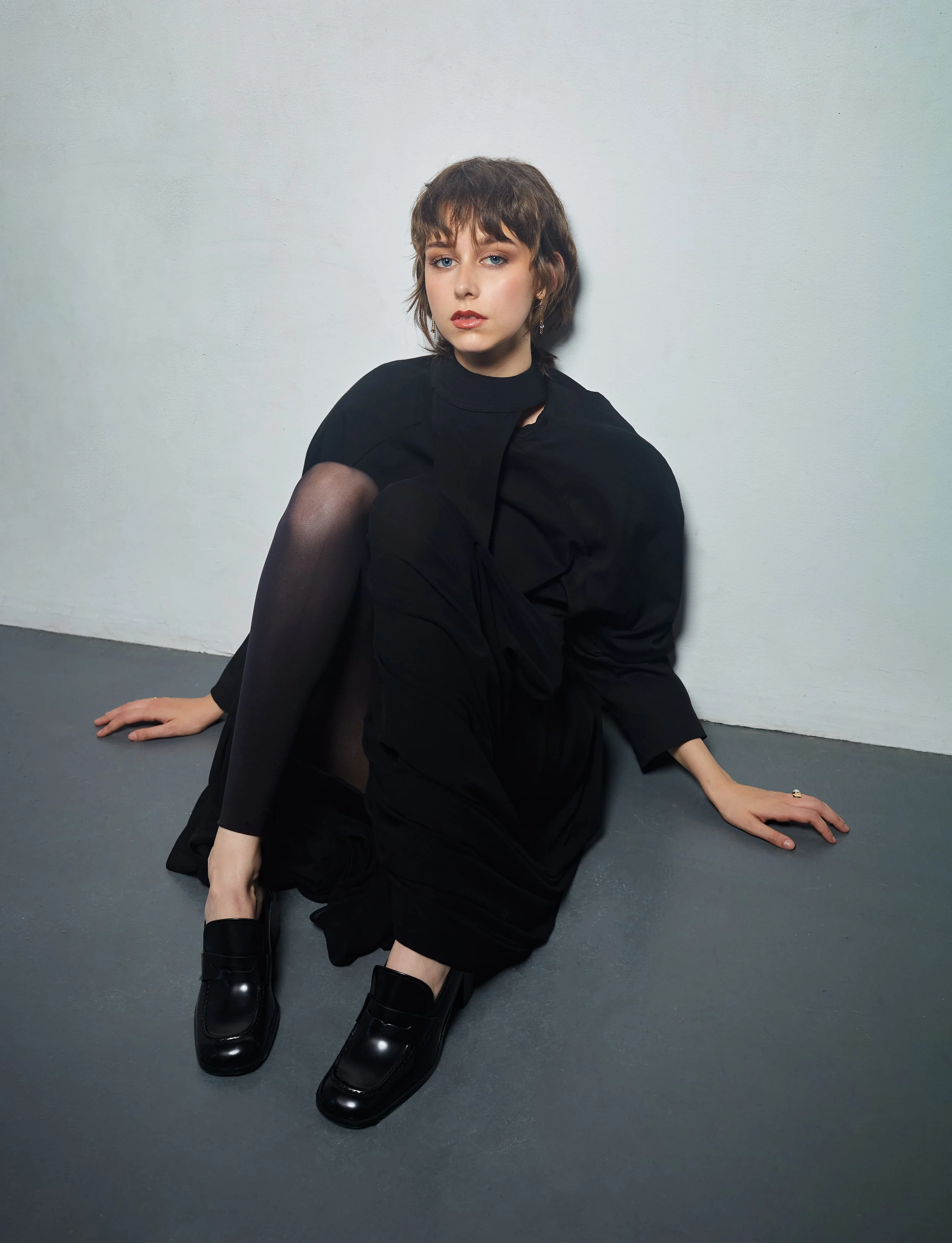 A woman with short brown hair and light skin sitting on the floor against a white wall, dressed in black, with one knee up and wearing black shoes.