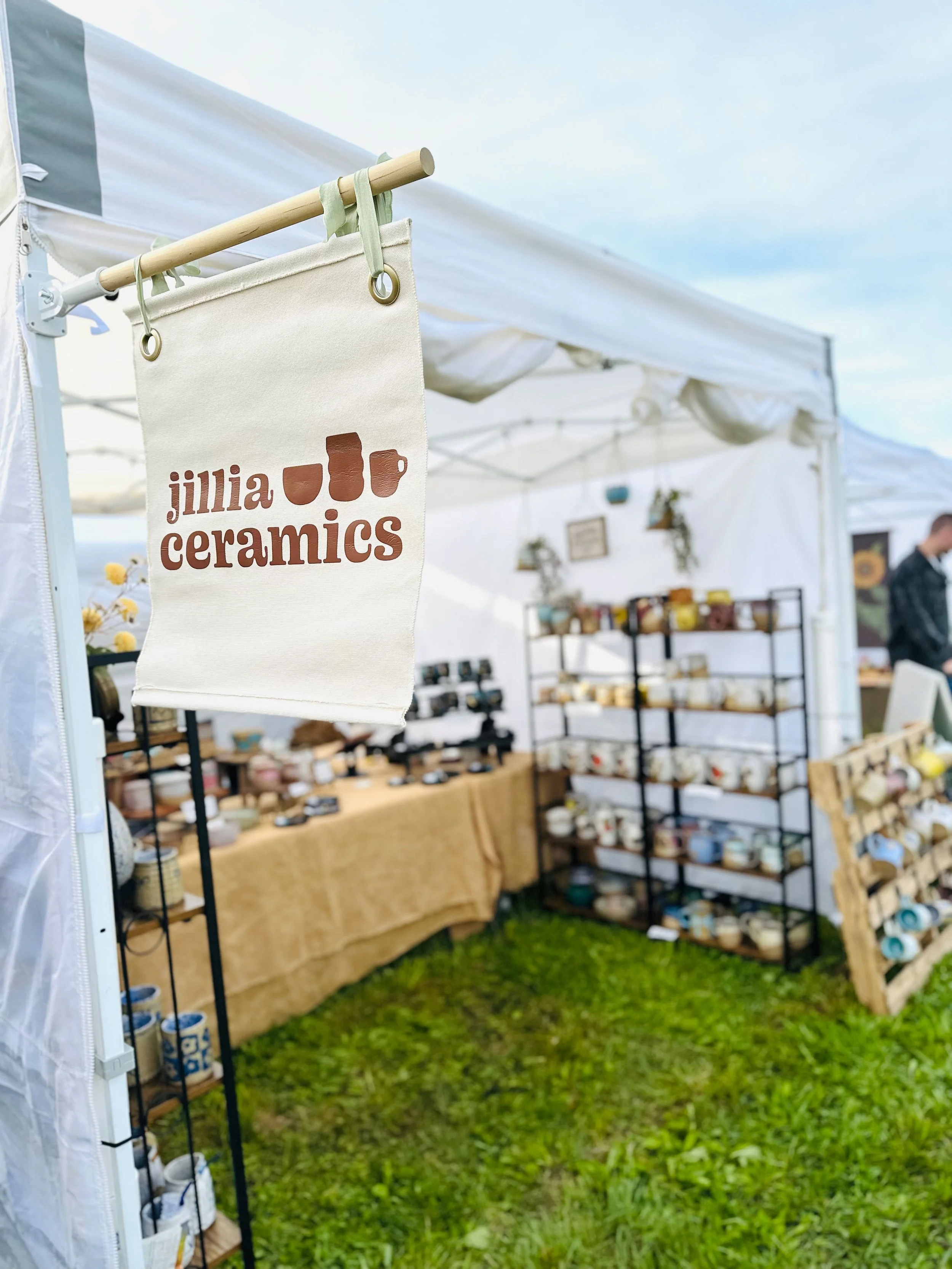 A tent with a sign that reads 'jillia ceramics' hanging from a wooden rod with ceramic mugs and pottery inside the tent, set up outdoors on green grass.