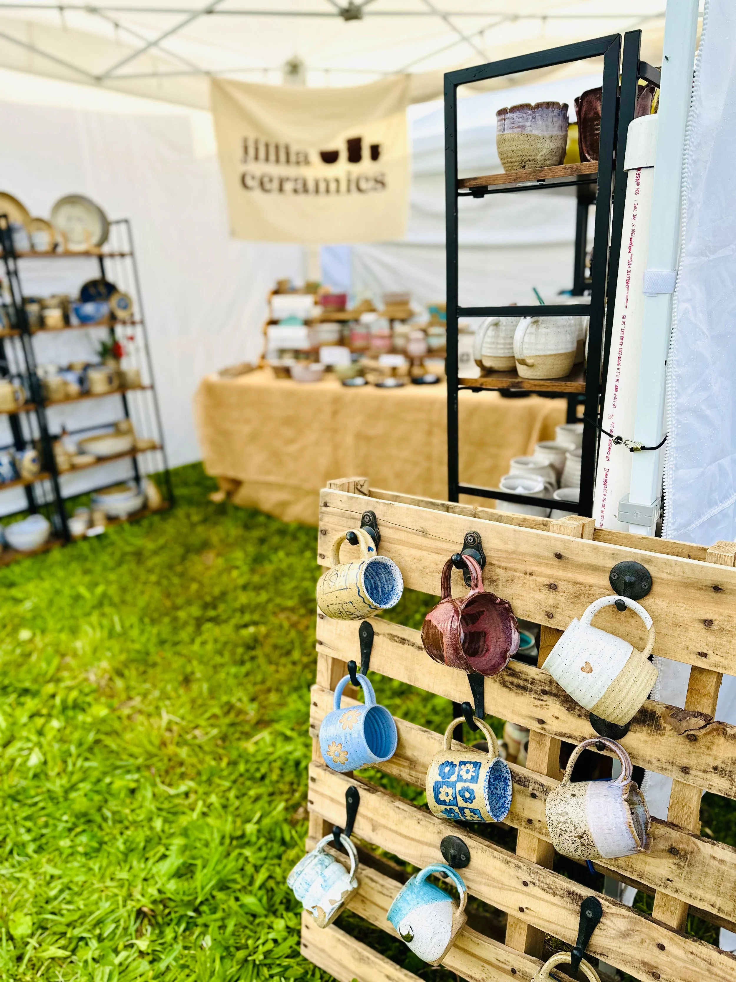 Wooden display stand with colorful ceramic and porcelain mugs hanging on hooks outdoors at a craft fair, with grass beneath and additional shelves of pottery in the background under a white canopy.
