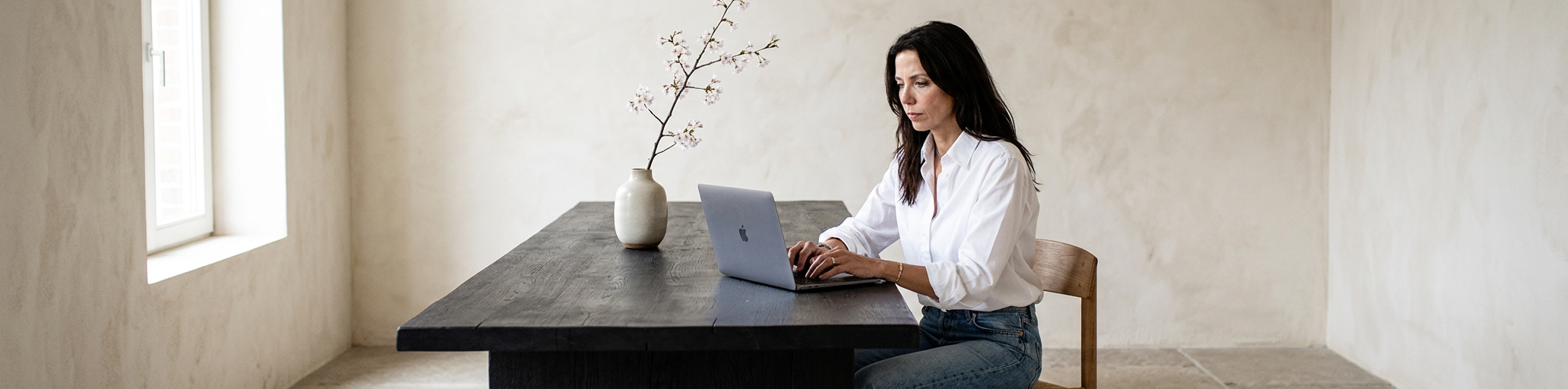 Frau arbeitet an einem Laptop in einem minimalistischen Raum mit weißer Wand und einer Vase mit blühendem Zweig auf dem Tisch.