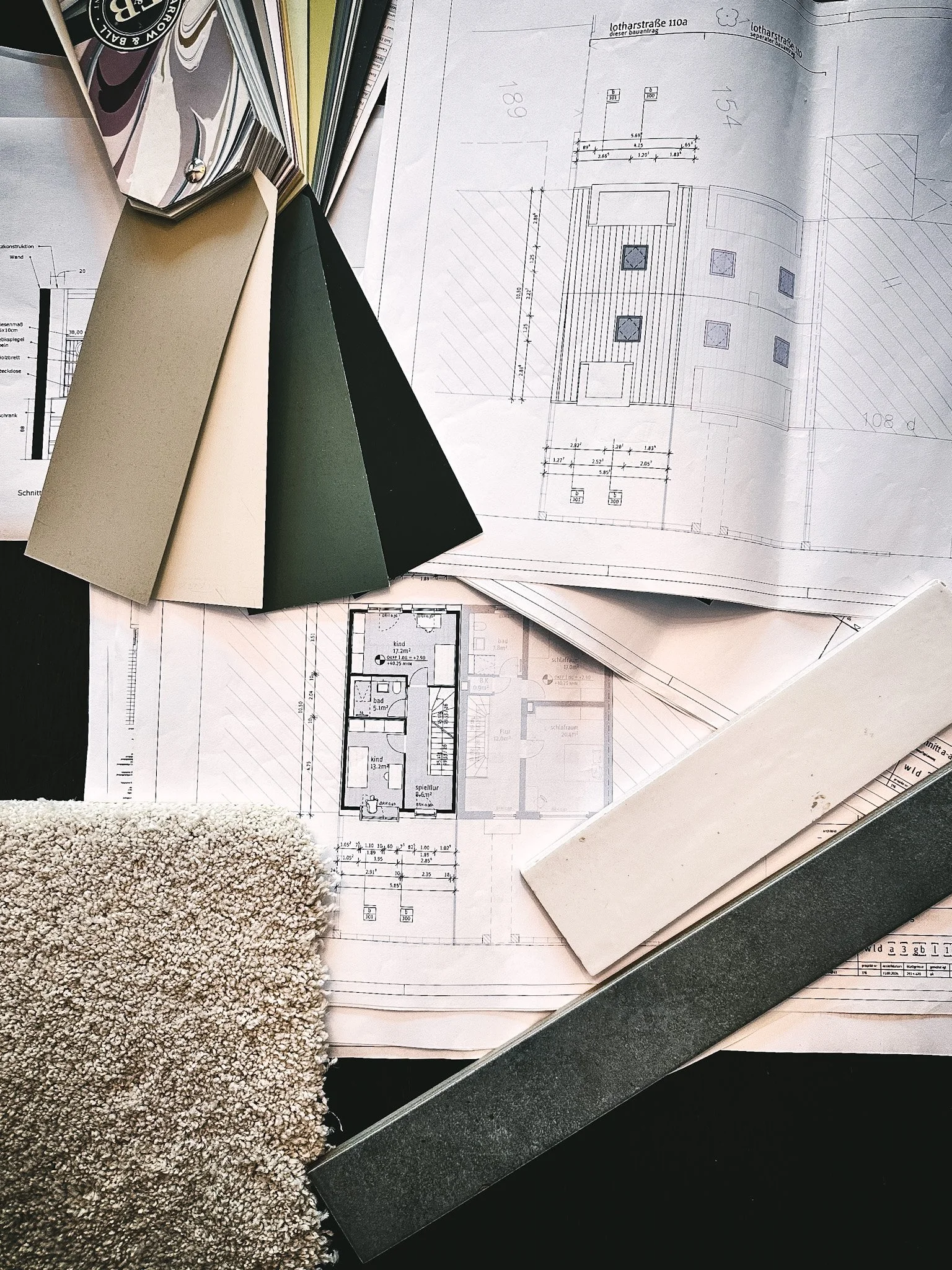 Architektenpläne, Farbproben und ein Teppich liegen auf einem Tisch, vermutlich in einem Architekturbüro oder Planungsraum.