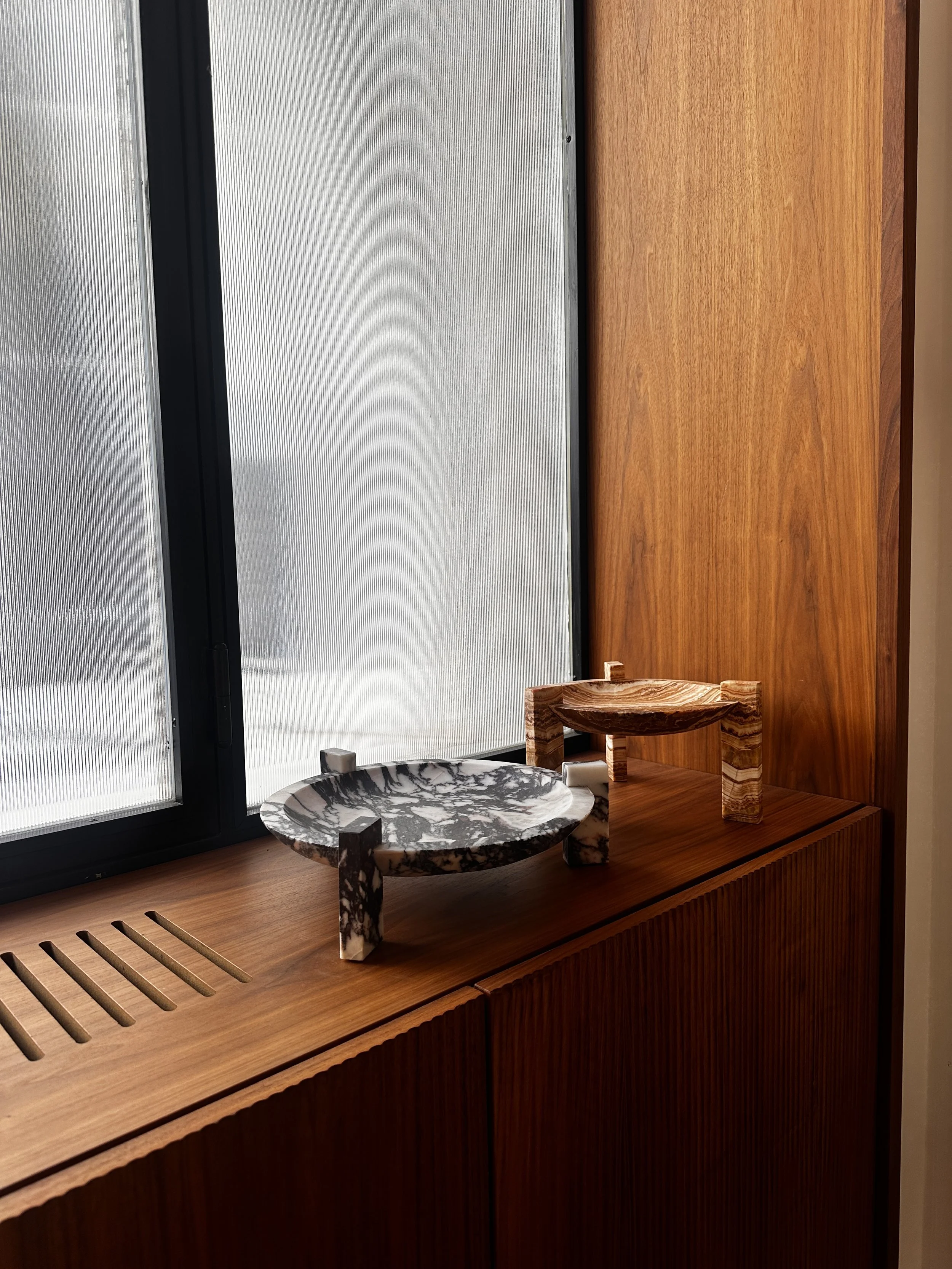 Ein Fenster mit halbtransparentem, strukturiertem Glas vor einem Holzregal mit dekorativen Gegenständen aus Marmor, darunter eine kleine Bühne und eine Schale.