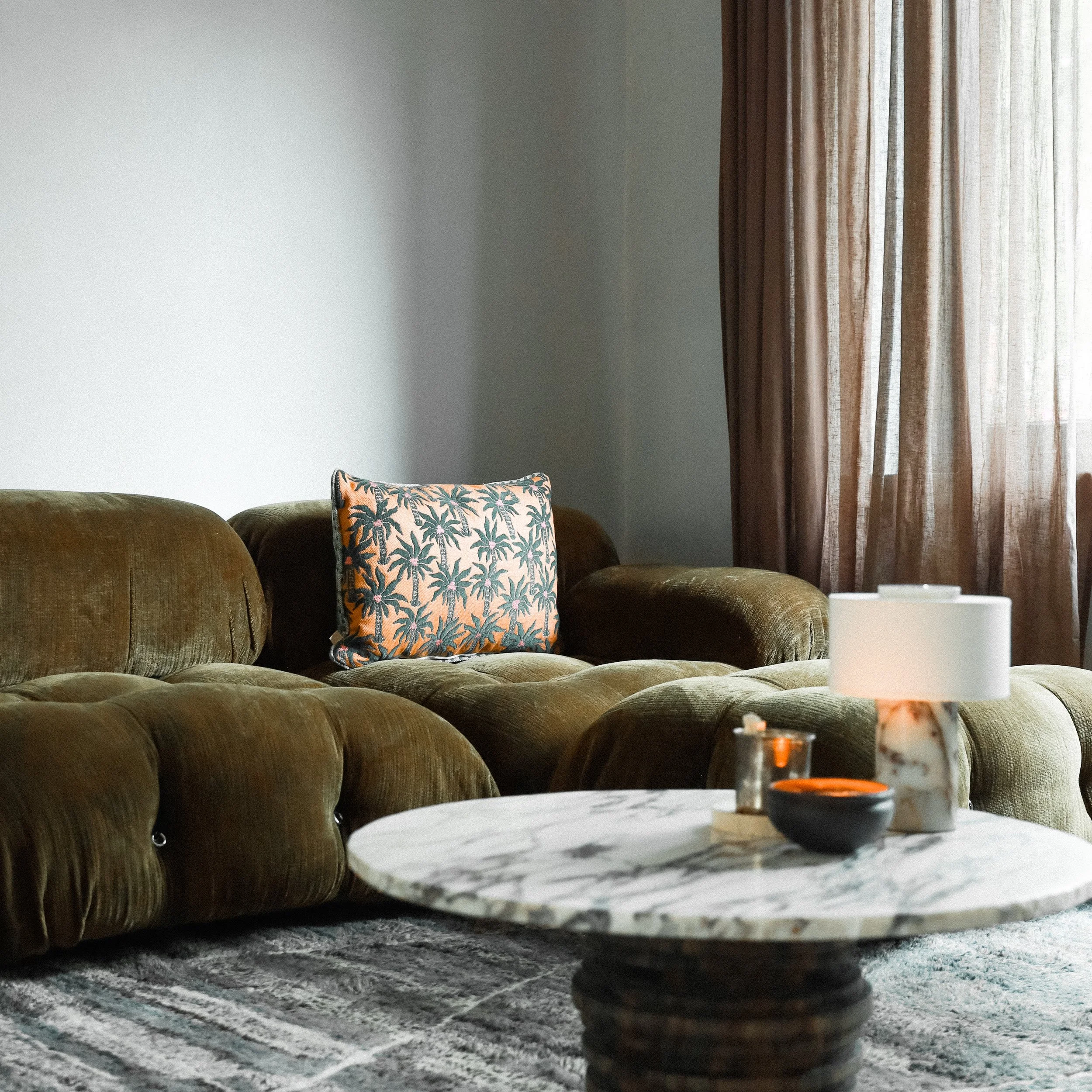 Wohnzimmer mit grüner Couch und Kissen, Kaffeetisch mit Kerze, Fenster mit lichtdurchlässigen Vorhängen.