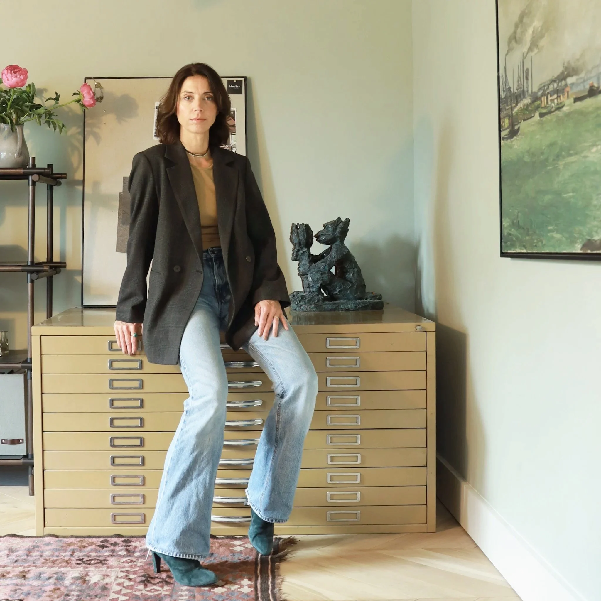 Frau steht vor einem Kasten mit Schubladen in einem Raum, trägt eine schwarze Blazer, Jeans und eine Halskette, im Hintergrund ein Gemälde und eine Vase mit pinken Blumen.