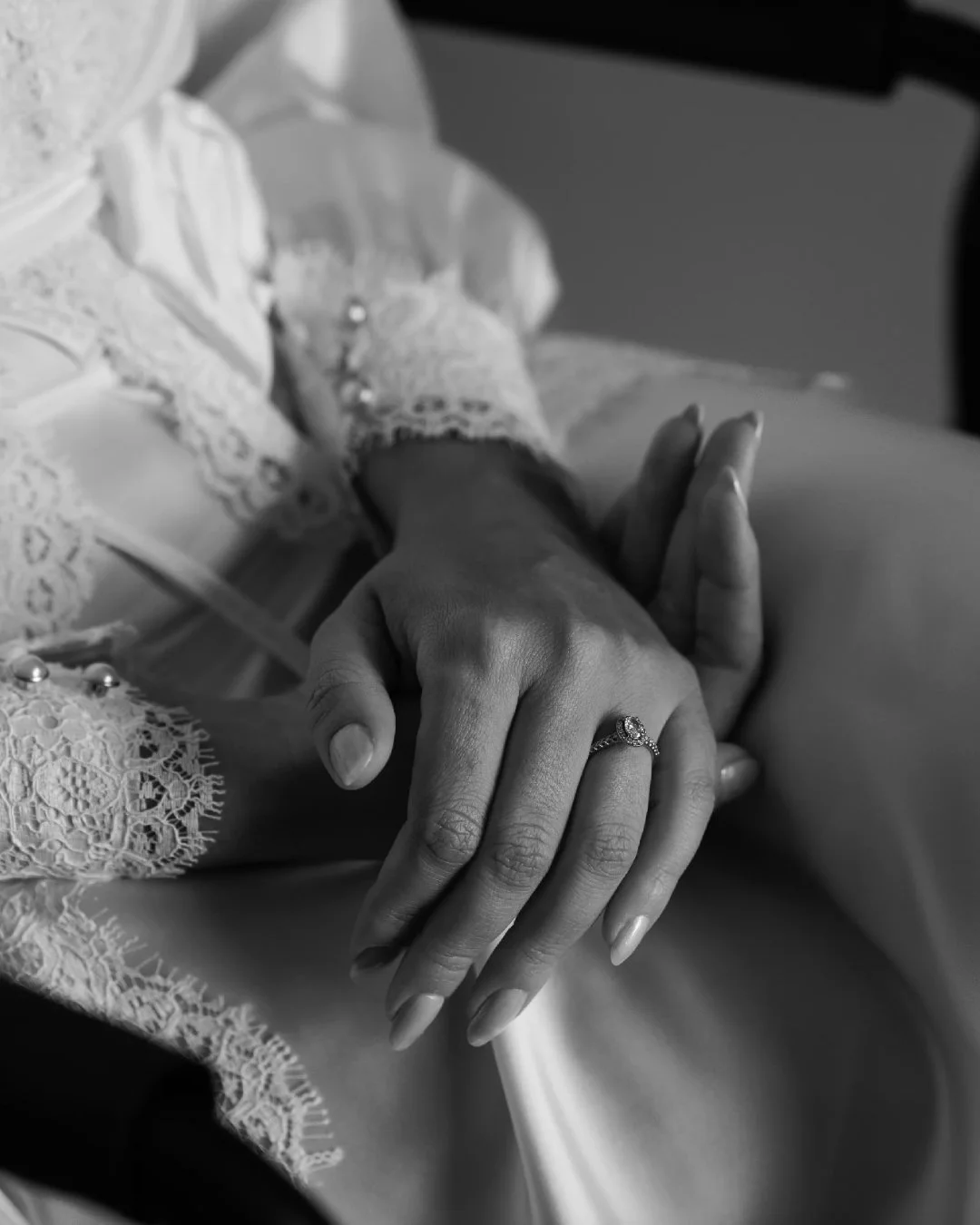 Close-up of a woman's hand with an engagement ring resting on her other hand, wearing lace clothing, near a lace-trimmed fabric.