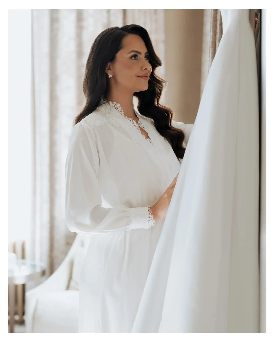 A woman in a white lace and satin nightgown standing by a window, smiling, with long dark hair and earrings, in a softly lit room.