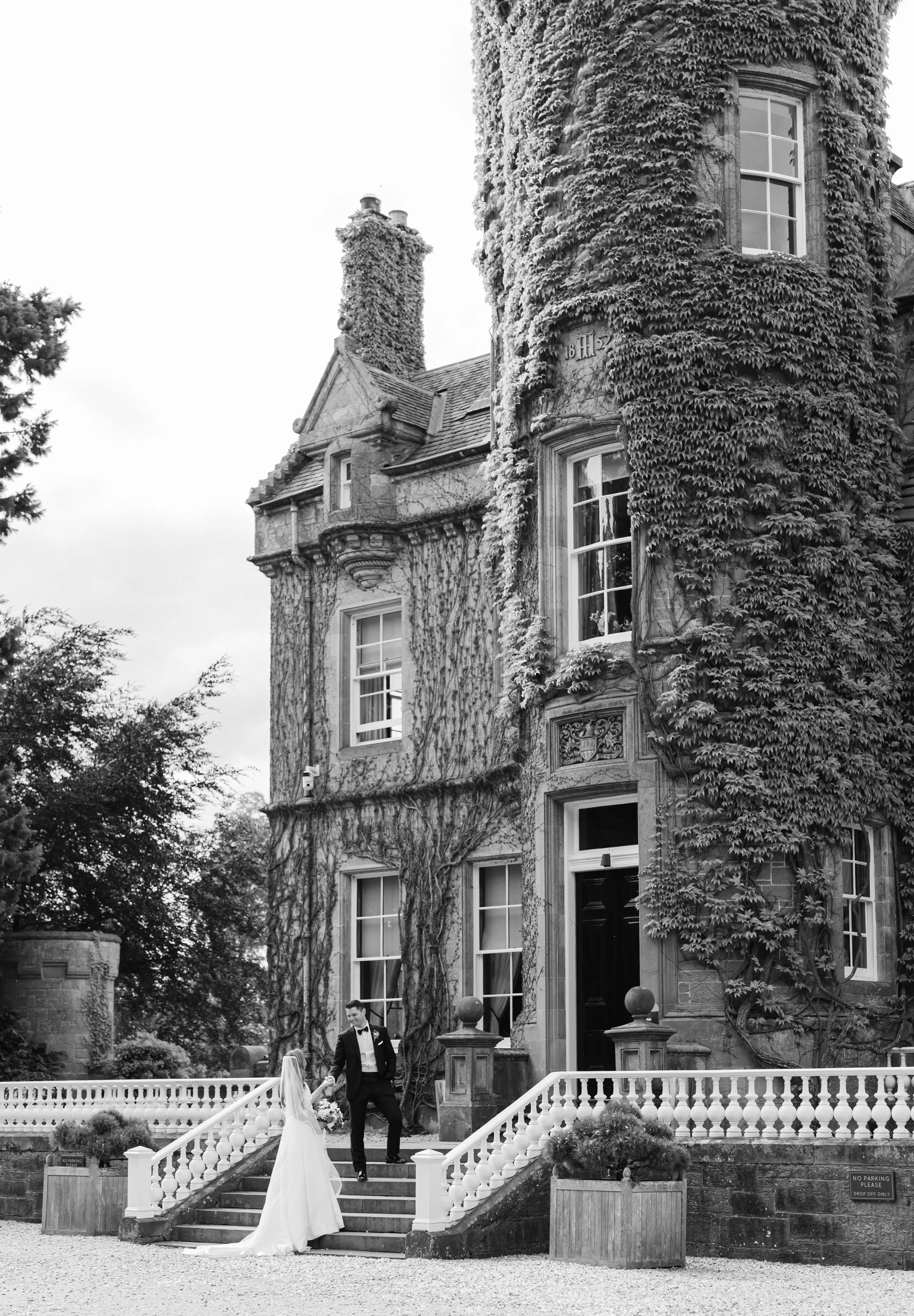 A bride and groom in wedding attire standing on steps outside a large, ivy-covered mansion.