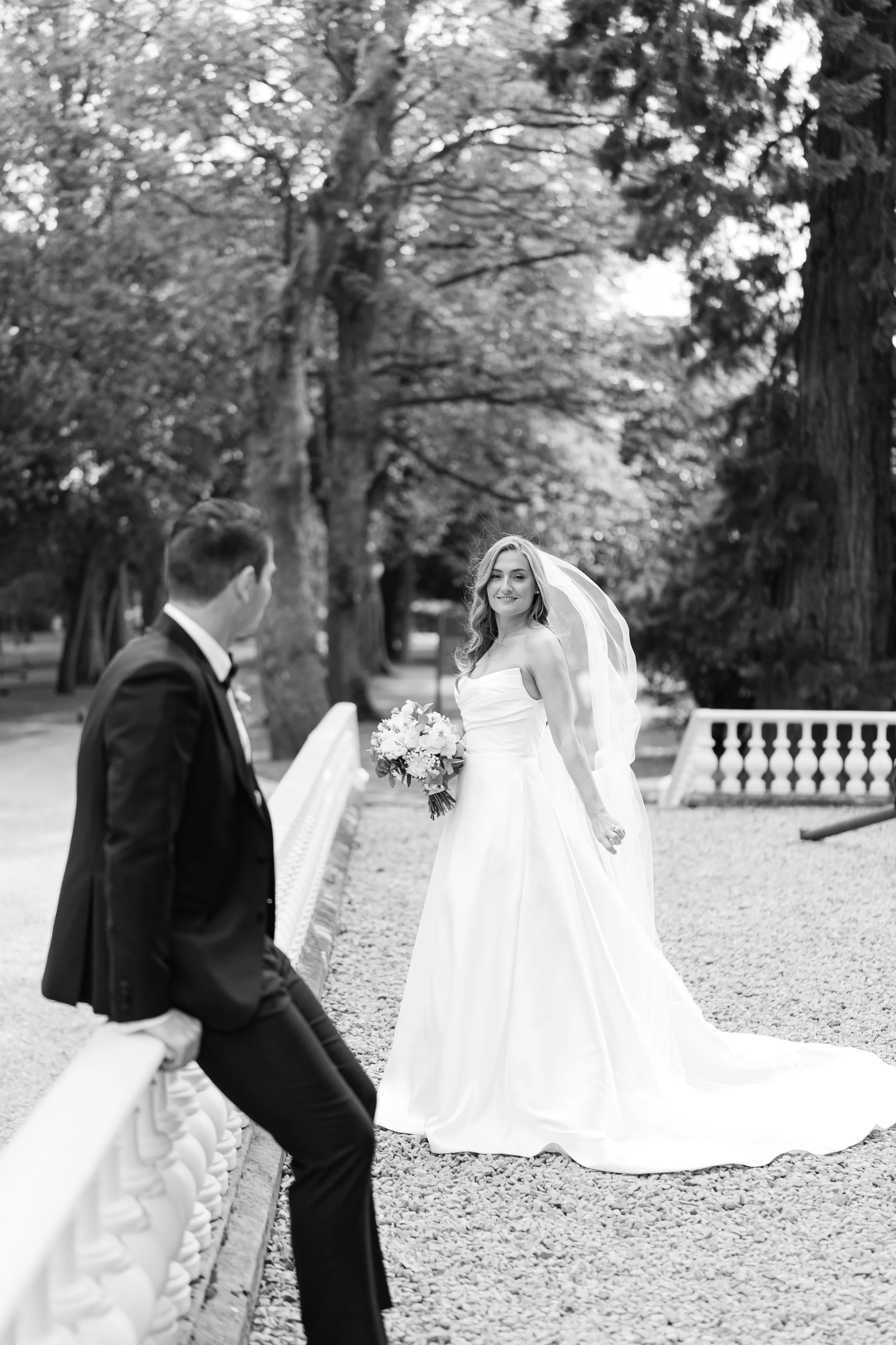A bride in a white wedding gown holding a bouquet, smiling, standing outdoors with a groom in a black suit sitting on a white fence, surrounded by trees.