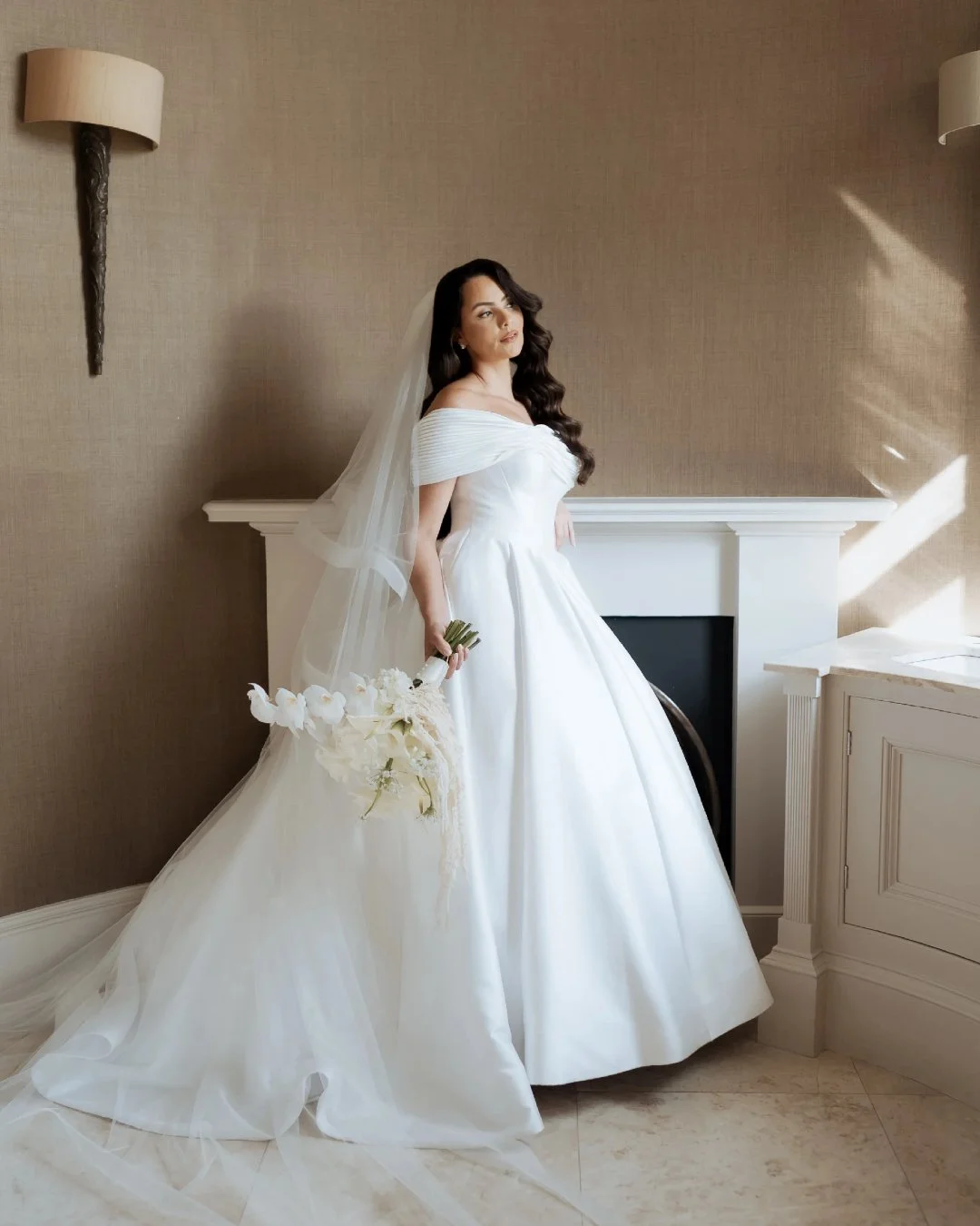 Bride in a white wedding dress holding a bouquet indoors near a fireplace.