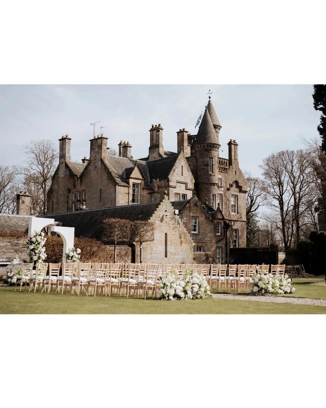 A historic castle-like building with a sloped roof and multiple chimneys, set against a backdrop of leafless trees. In the foreground, there are rows of chairs arranged for an outdoor wedding ceremony, decorated with white flowers and greenery.