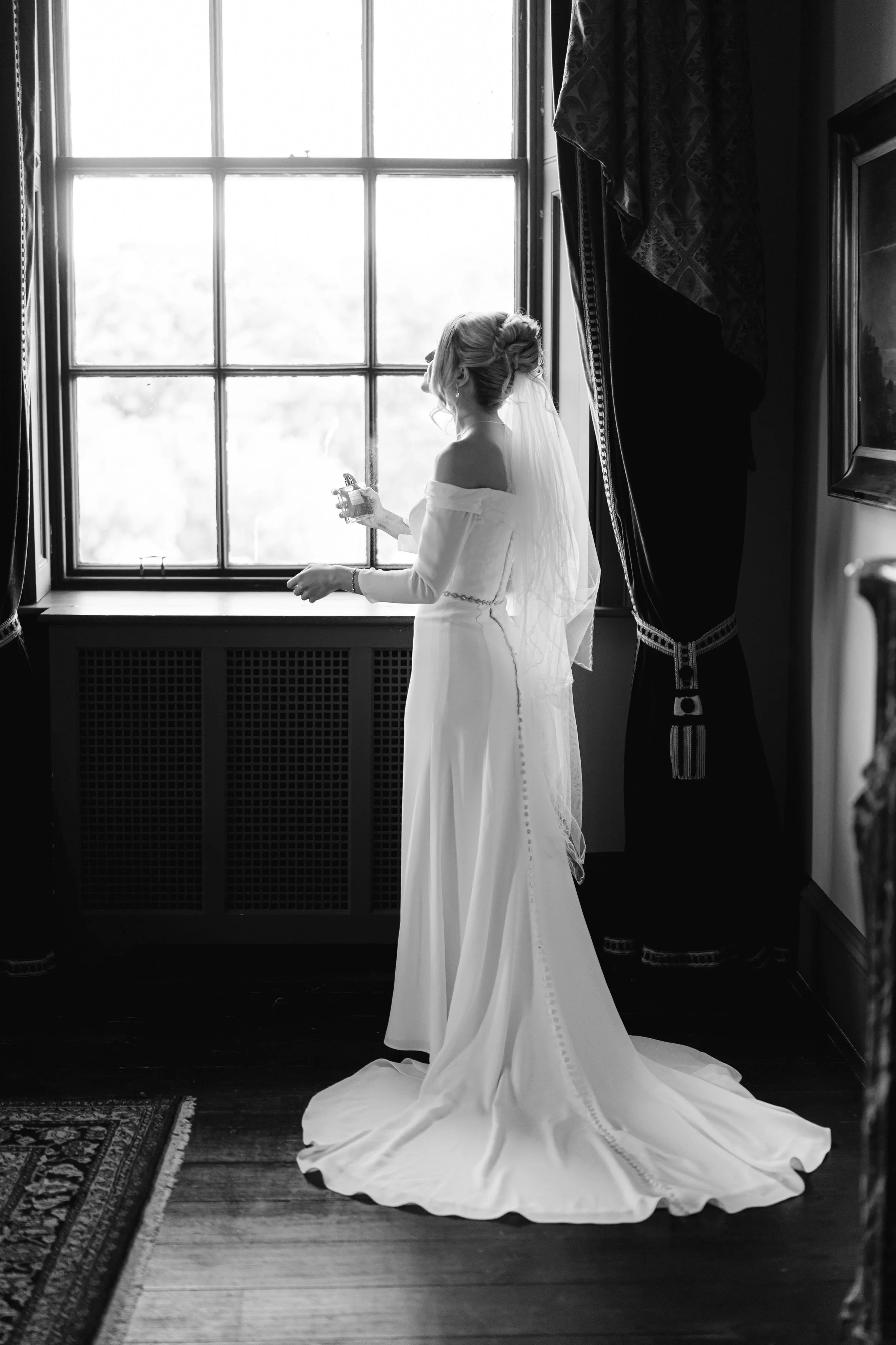A bride in a wedding dress stands by a window, holding a small bottle, with her face turned away from the camera in black and white.