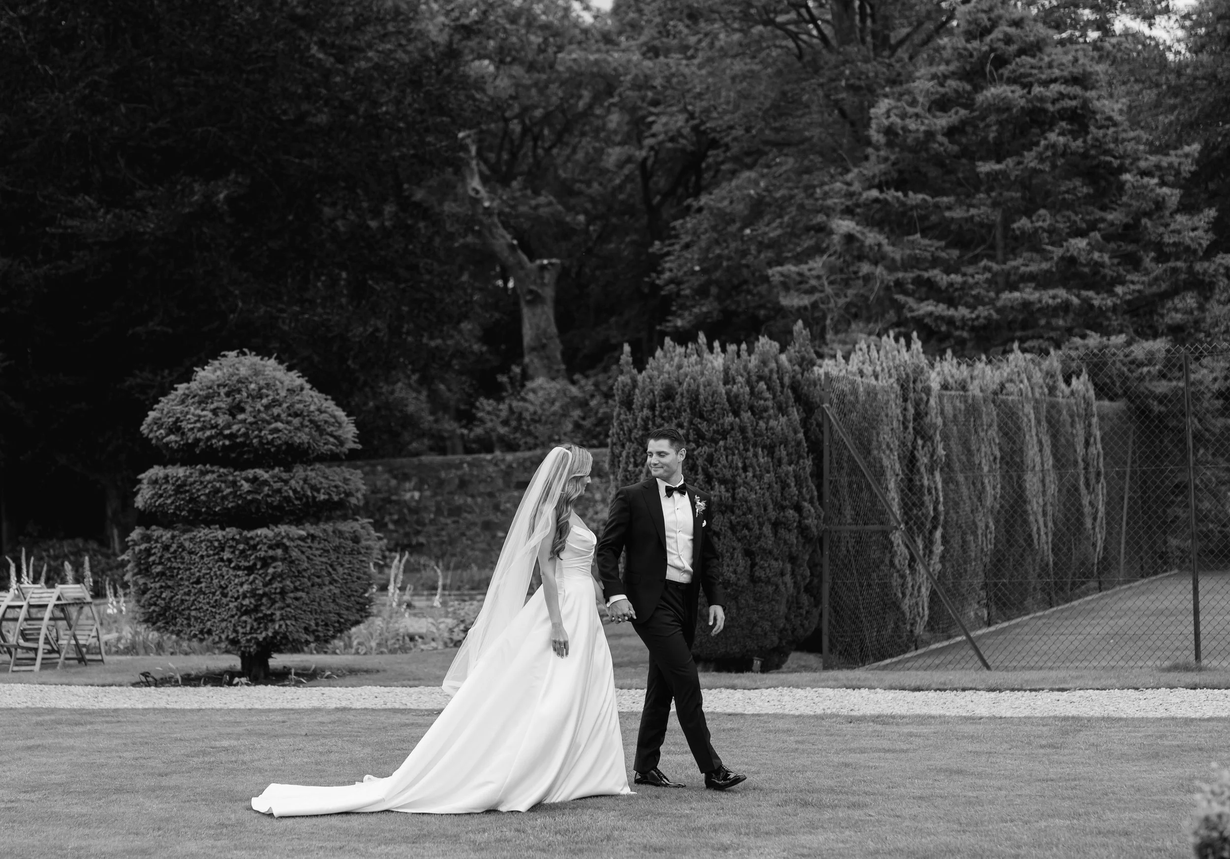 A bride and groom walking hand in hand on a lawn during their wedding. The bride is in a long white gown with a veil, and the groom is in a black tuxedo. There are trees and hedges in the background.