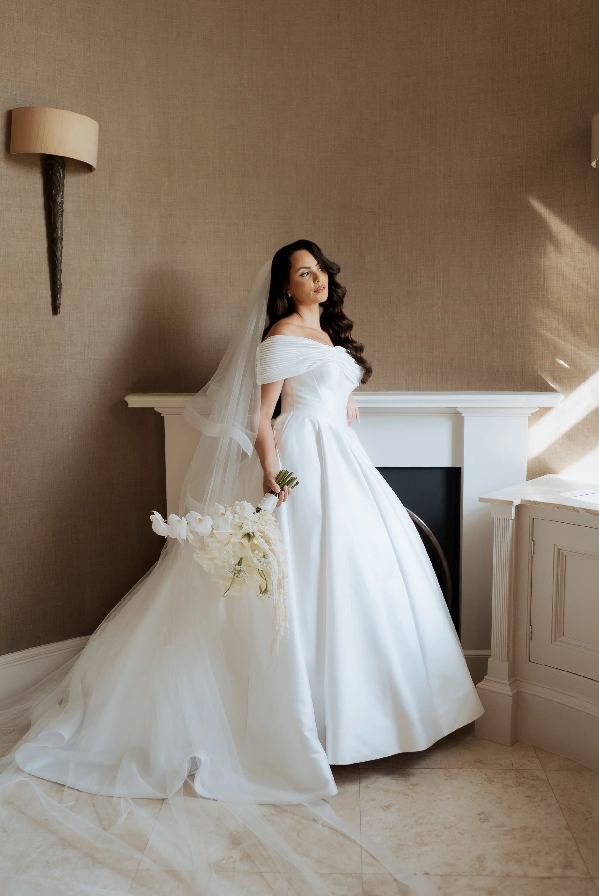 A bride with long dark hair in a white wedding gown and veil holding a bouquet of white flowers, standing indoors by a wall and fireplace.