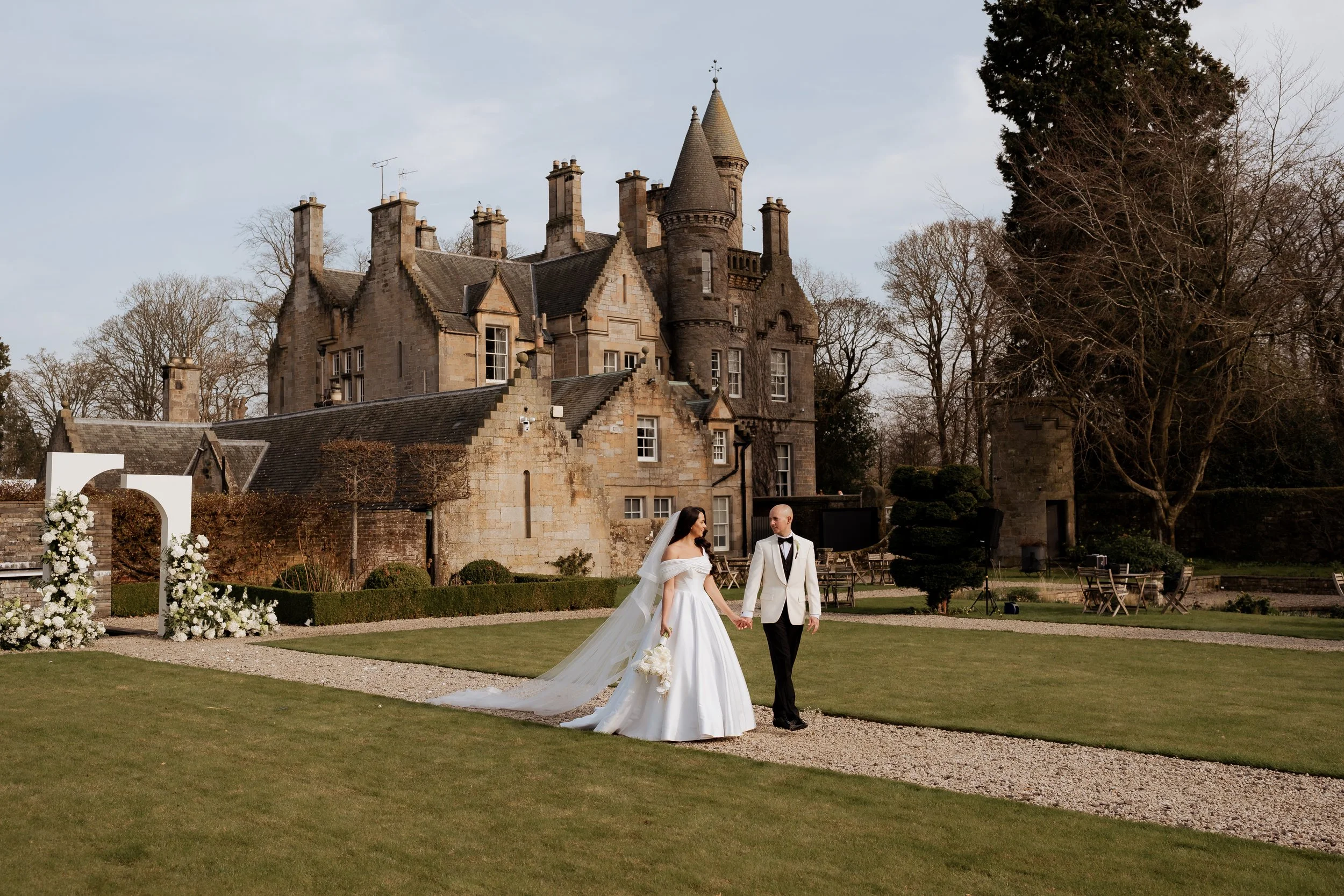 A bride and groom holding hands walking on a gravel path in front of a castle-like building, surrounded by trees and outdoor tables, on a cloudy day.