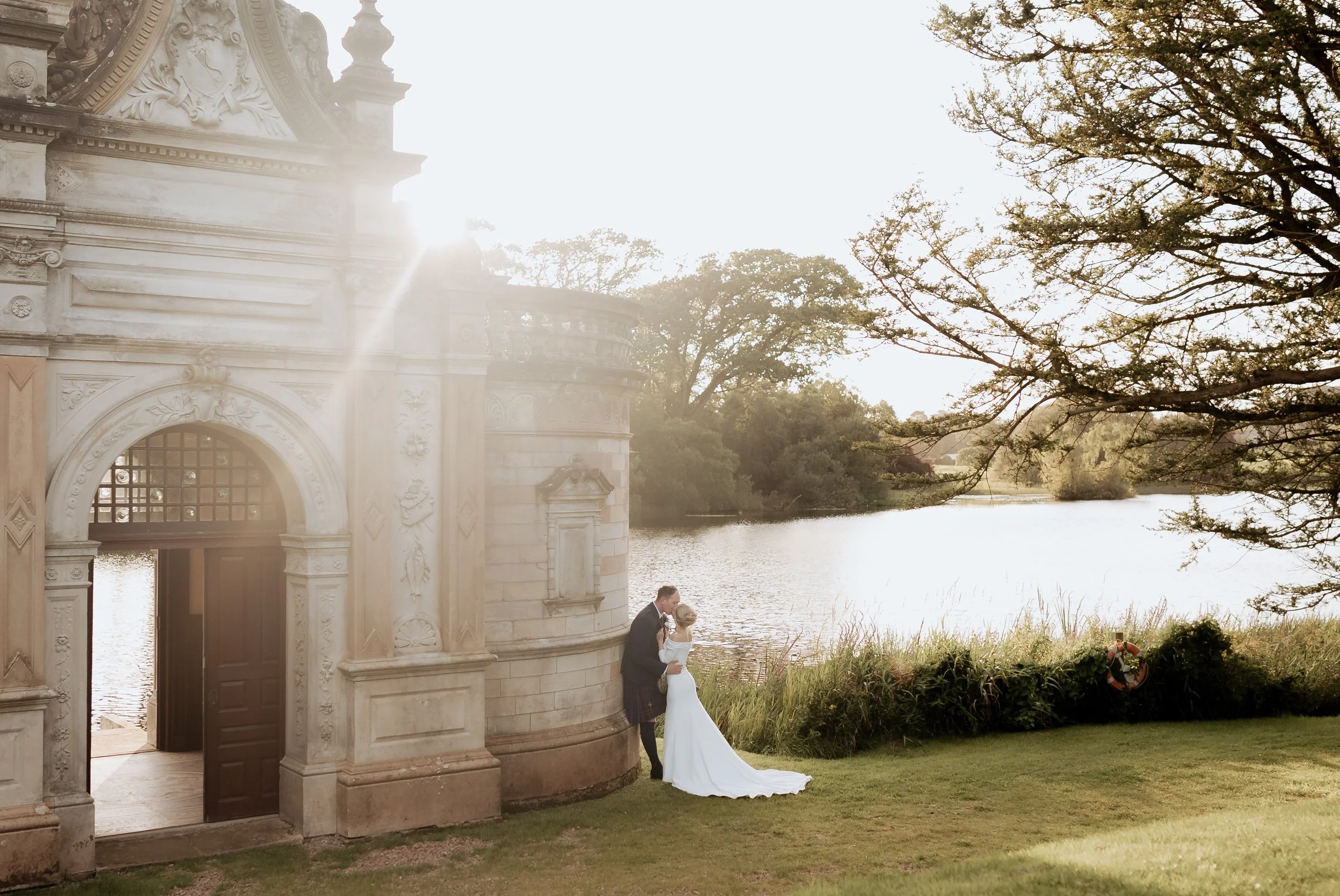 A bride and groom kiss near a historic building next to a river, with trees and grasses in the background, during sunset.