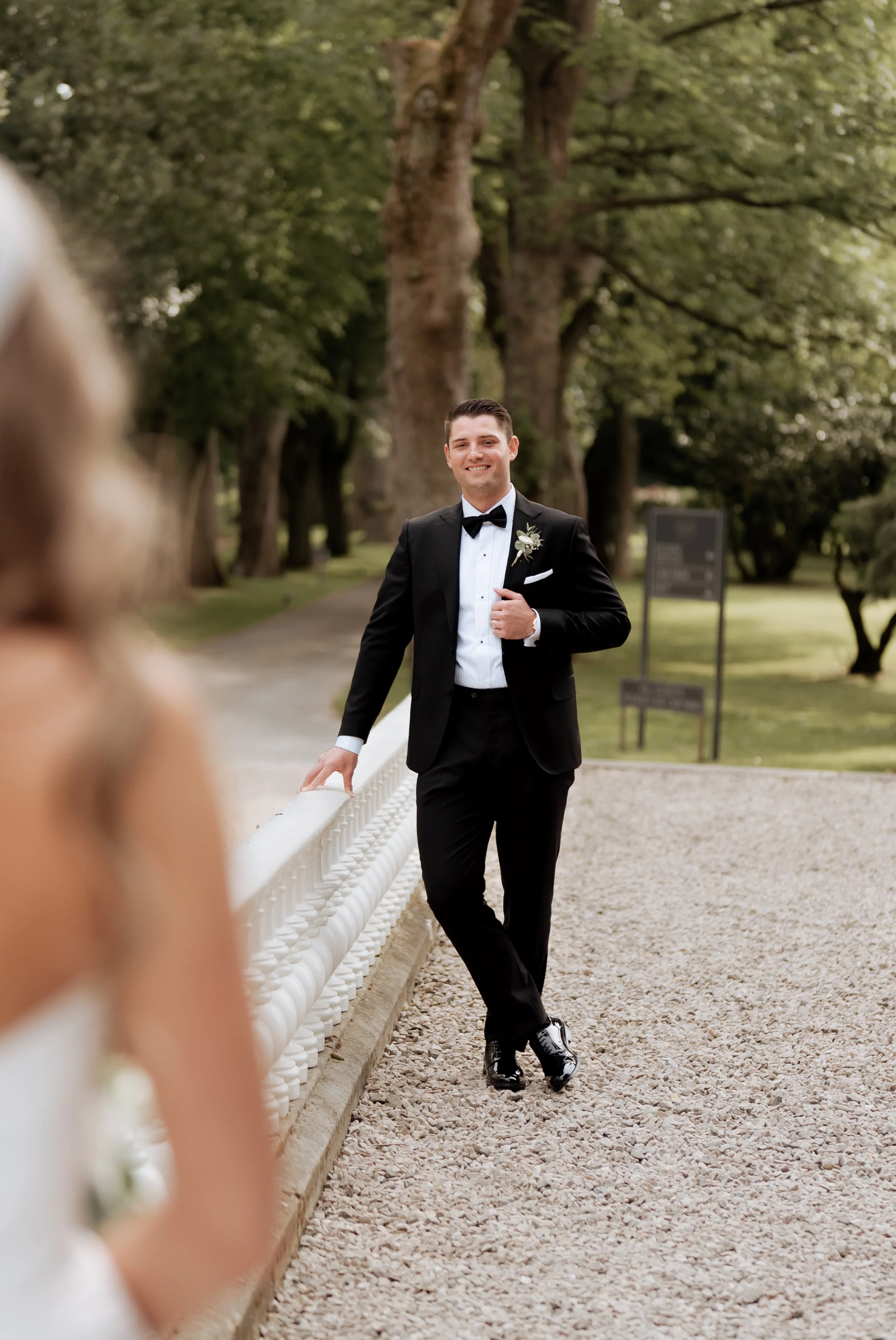 A groom in a black tuxedo and bow tie, smiling, standing outdoors next to a white railing in a park-like setting with trees and grass.