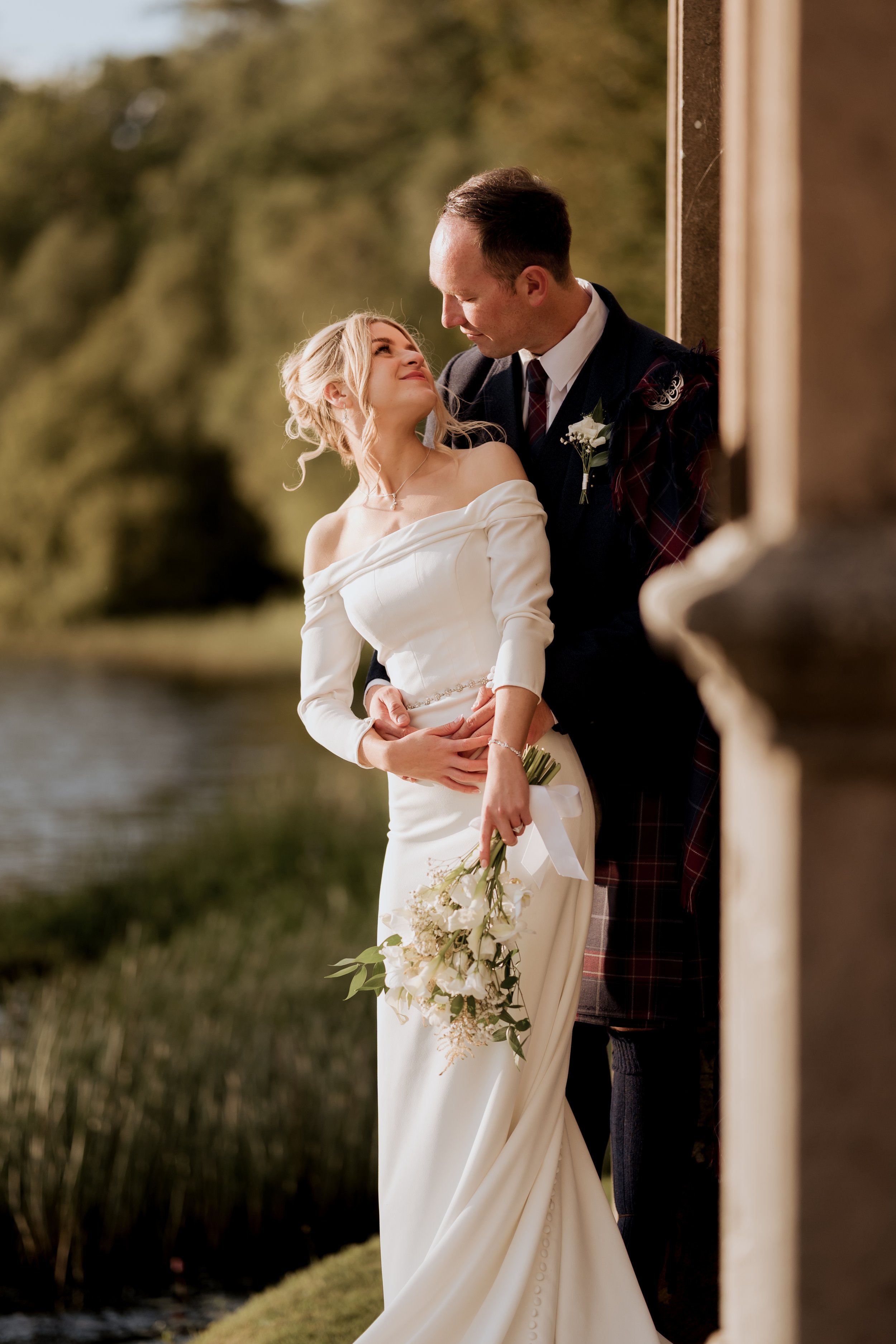 A bride and groom embrace outdoors near a body of water, with trees in the background. The bride is holding a bouquet of white flowers and wearing an off-shoulder white wedding dress. The groom is leaning on a stone structure, wearing a dark suit wit