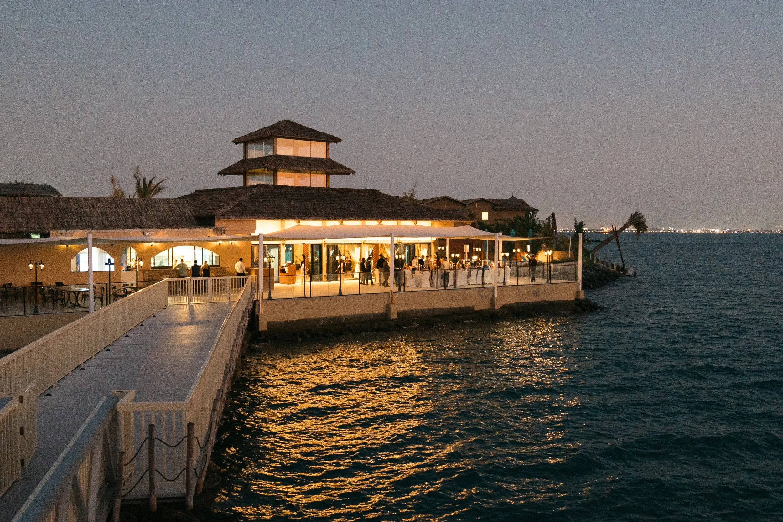 An illuminated seaside restaurant at night with people dining outside, a wooden pier extending into the water, and a city skyline in the distance.