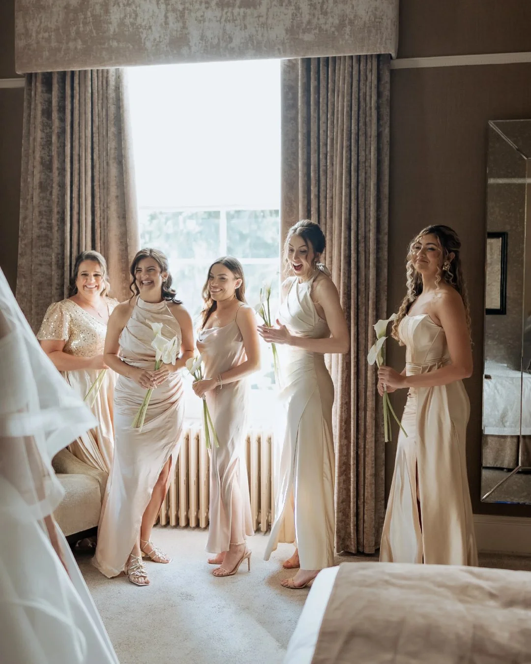 A group of six women in elegant cream-colored dresses standing in a hotel room near a window, holding white calla lilies, smiling and celebrating together.