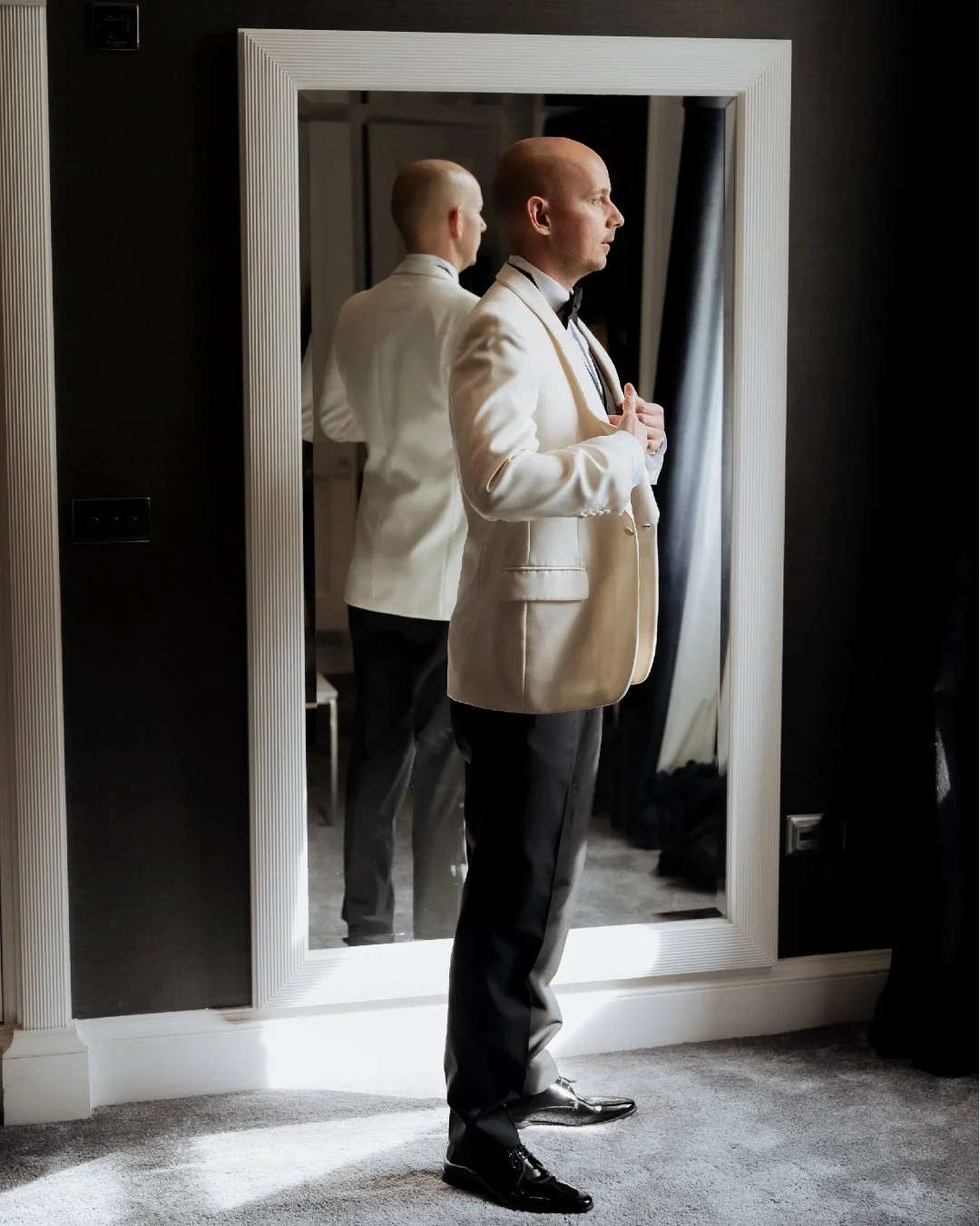 Man dressed in a beige tuxedo jacket and dark pants, standing in front of a mirror, adjusting his jacket, with a reflection visible in the mirror.