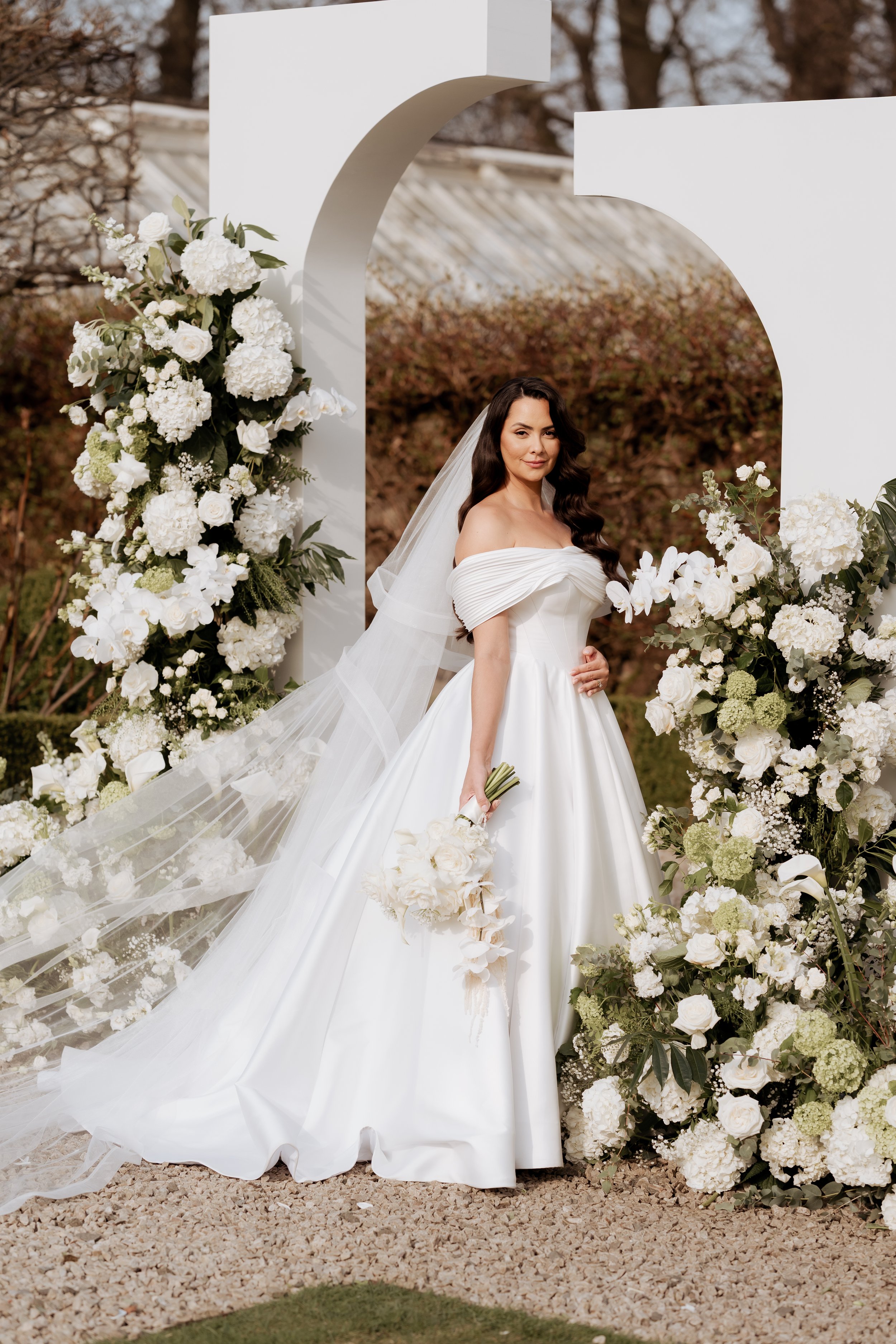 A bride in a white wedding gown holding a bouquet, standing under a large white arch decorated with white flowers, outdoors with trees and a rustic building in the background.