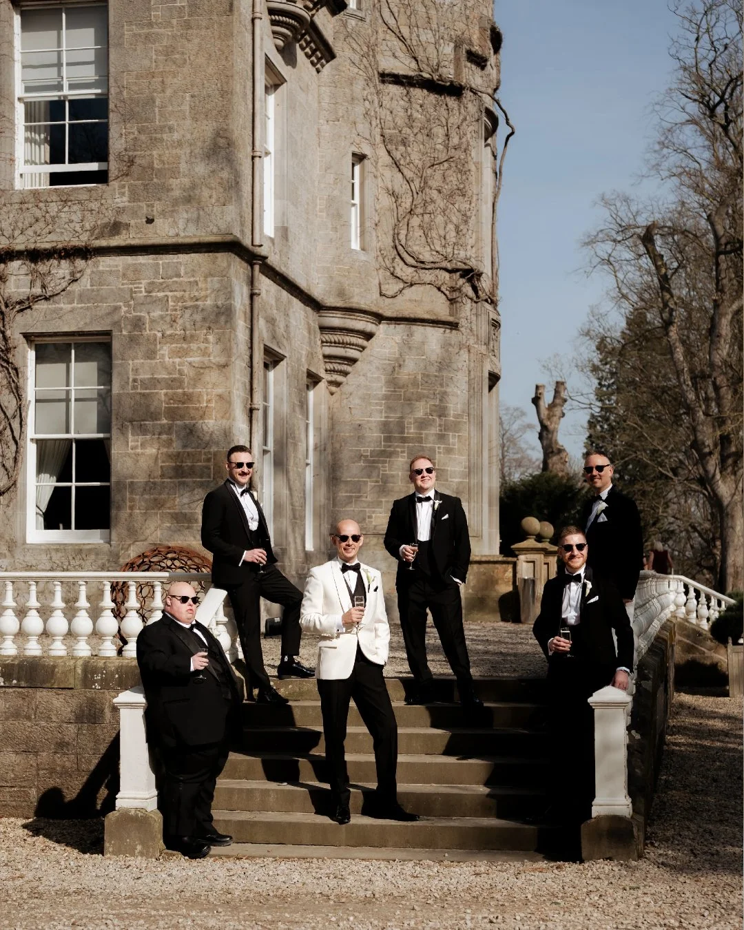 A group of six men dressed in tuxedos, one in a white tuxedo, standing on the steps of a historic stone building with large windows and a white railing. They are holding drinks and wearing sunglasses, smiling outdoors on a sunny day.