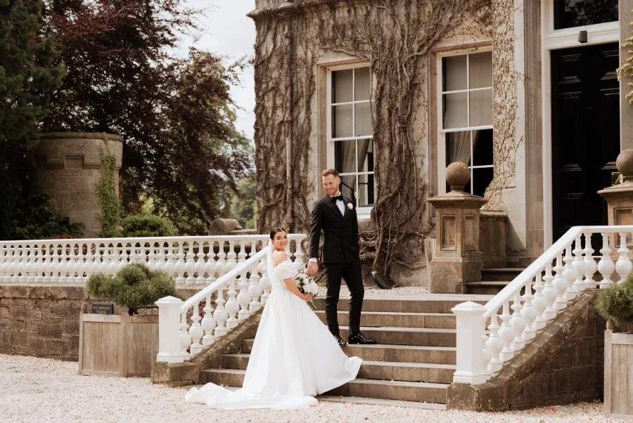 Bride and groom standing on front steps of a large historic house with a tree growing on the wall behind them, bride in a white wedding gown holding a bouquet, groom in a black suit and tie