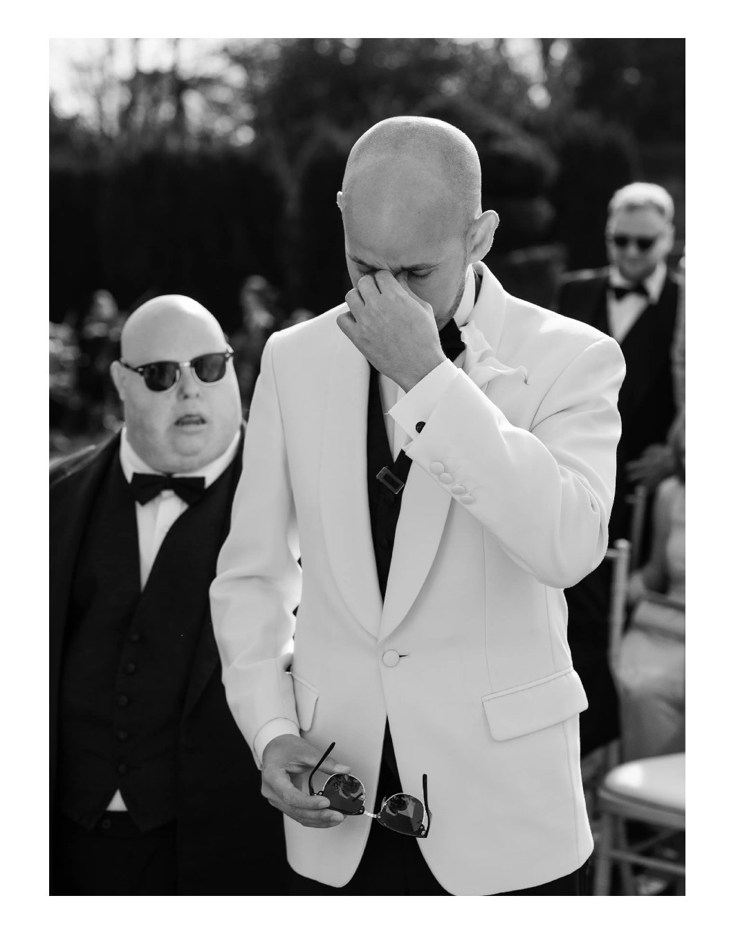 A man in a white tuxedo with a black shirt and tie, standing outdoors, holding sunglasses, and facepalming with his hand covering his face. Two men in black tuxedos and sunglasses are in the background.