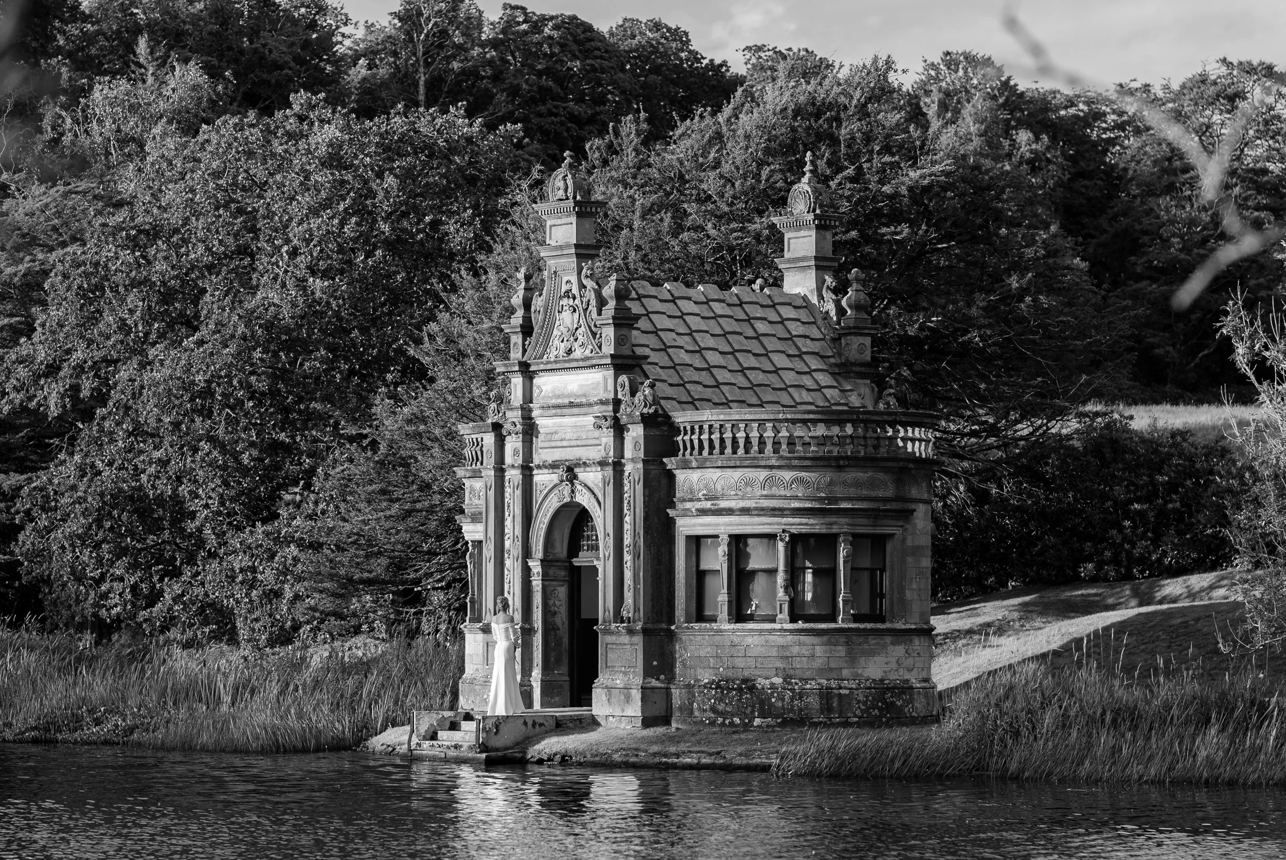 Black and white photograph of a small ornate building by a lake, surrounded by trees. A woman in a long dress stands on the steps near the entrance.