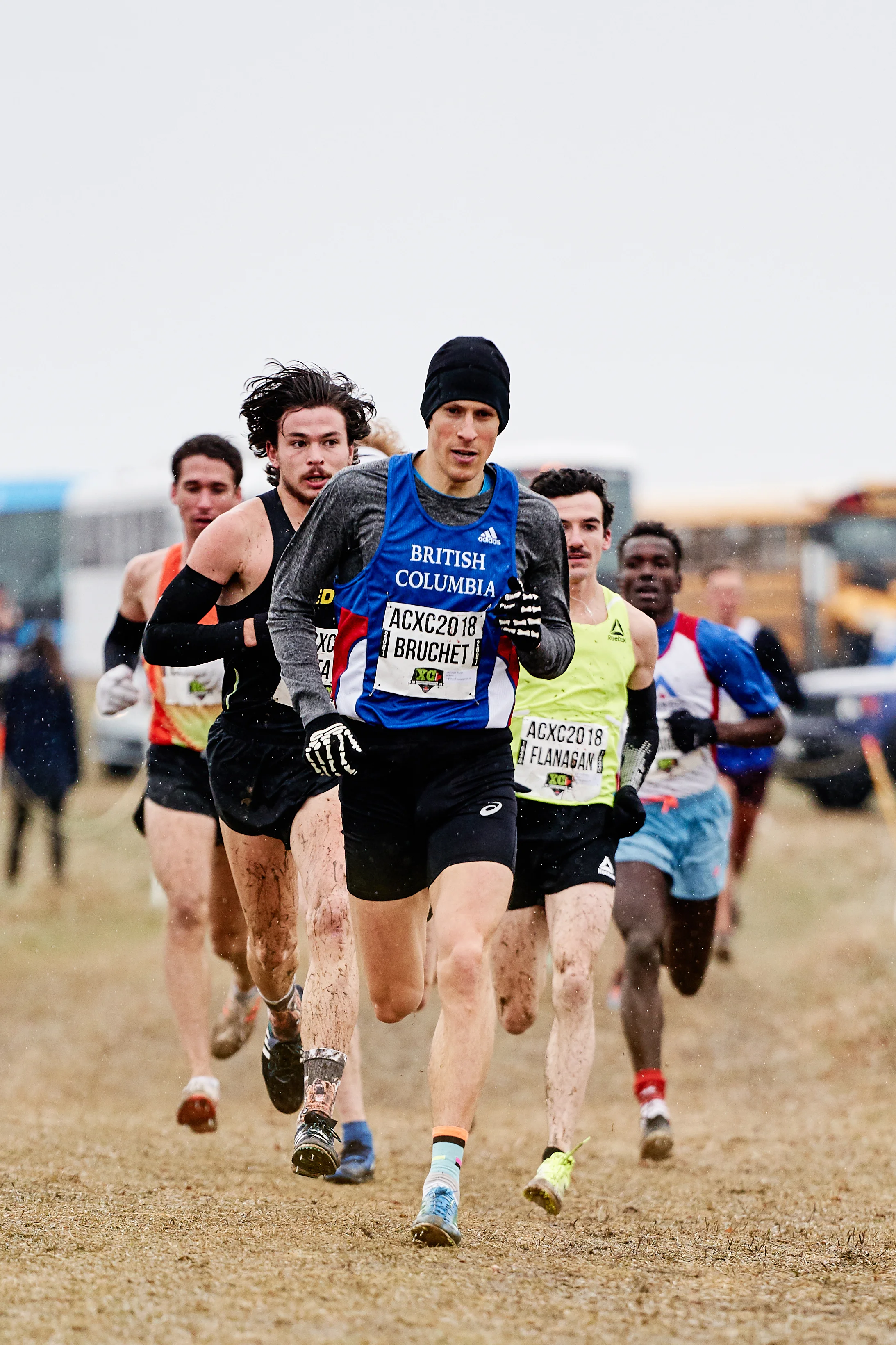Group of male athletes running in a cross-country race on a muddy field, wearing race bibs and athletic gear in cold weather.