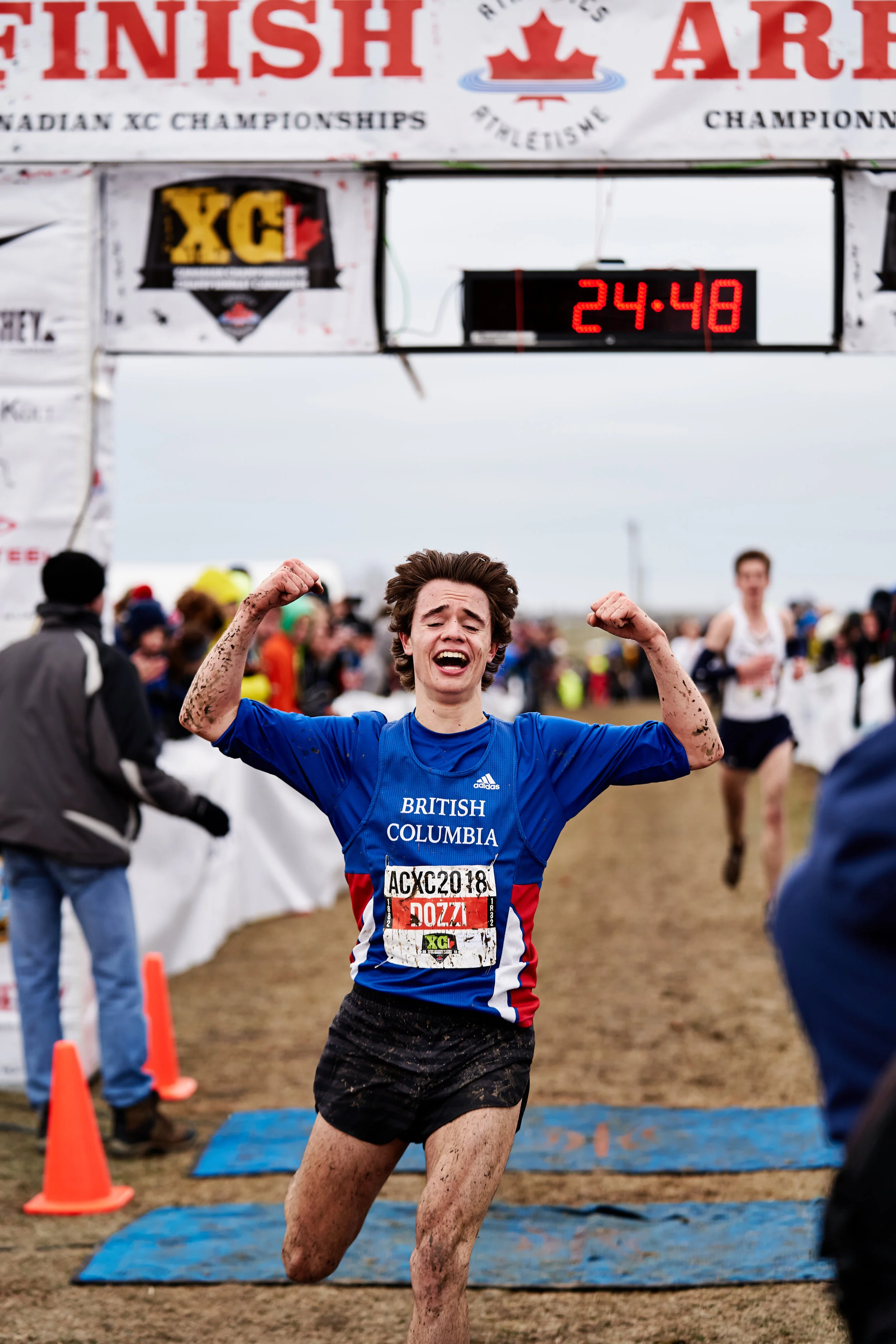 Excited young male athlete with mud on his arms and legs crossing the finish line of a race, celebrating his victory, wearing a blue shirt that reads "BRITISH COLUMBIA". The digital clock shows 24:48, and there are spectators and fellow runners in th