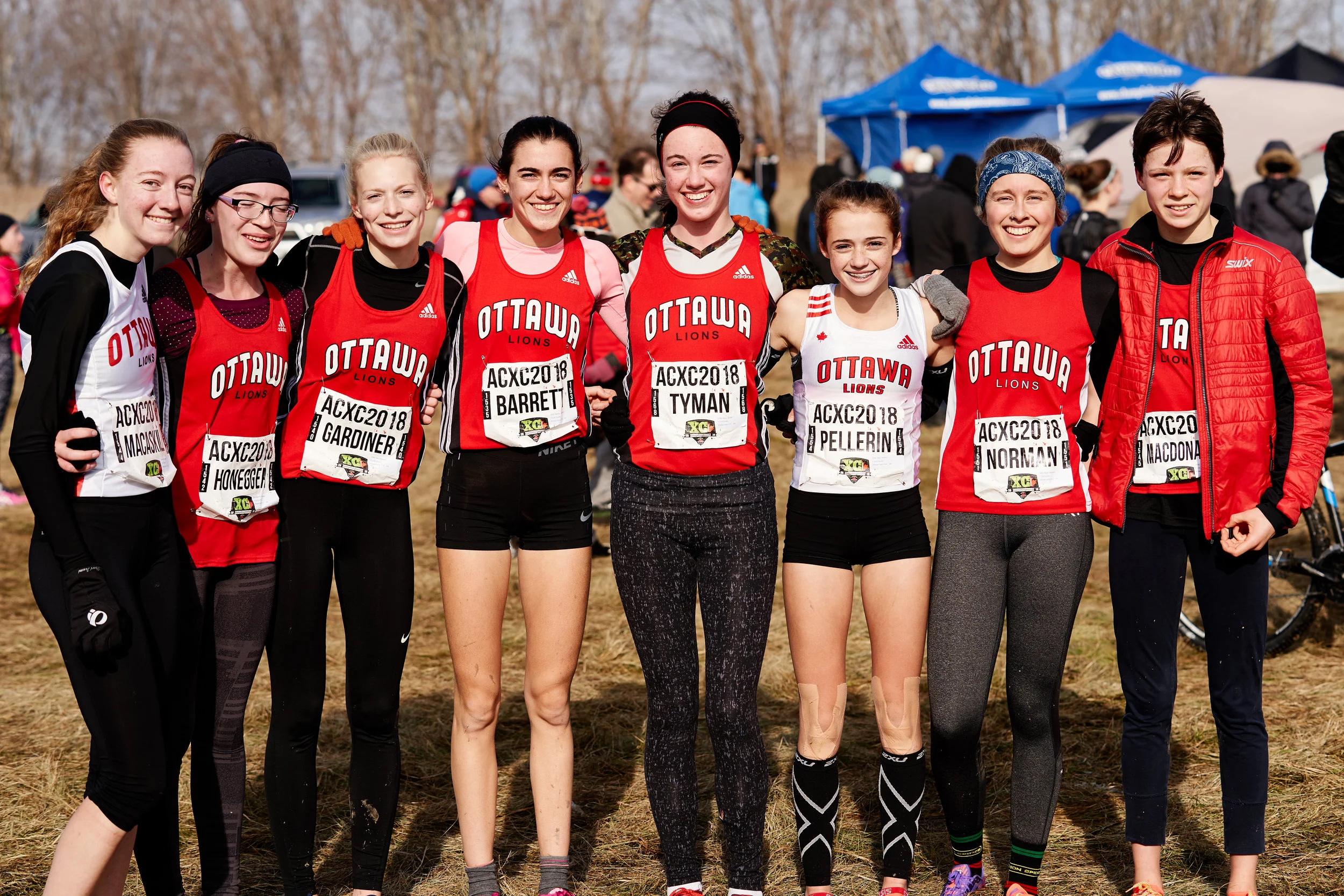Group of female cross-country runners in Ottawa Lions team uniforms standing outdoors at a race event, smiling with arms around each other.