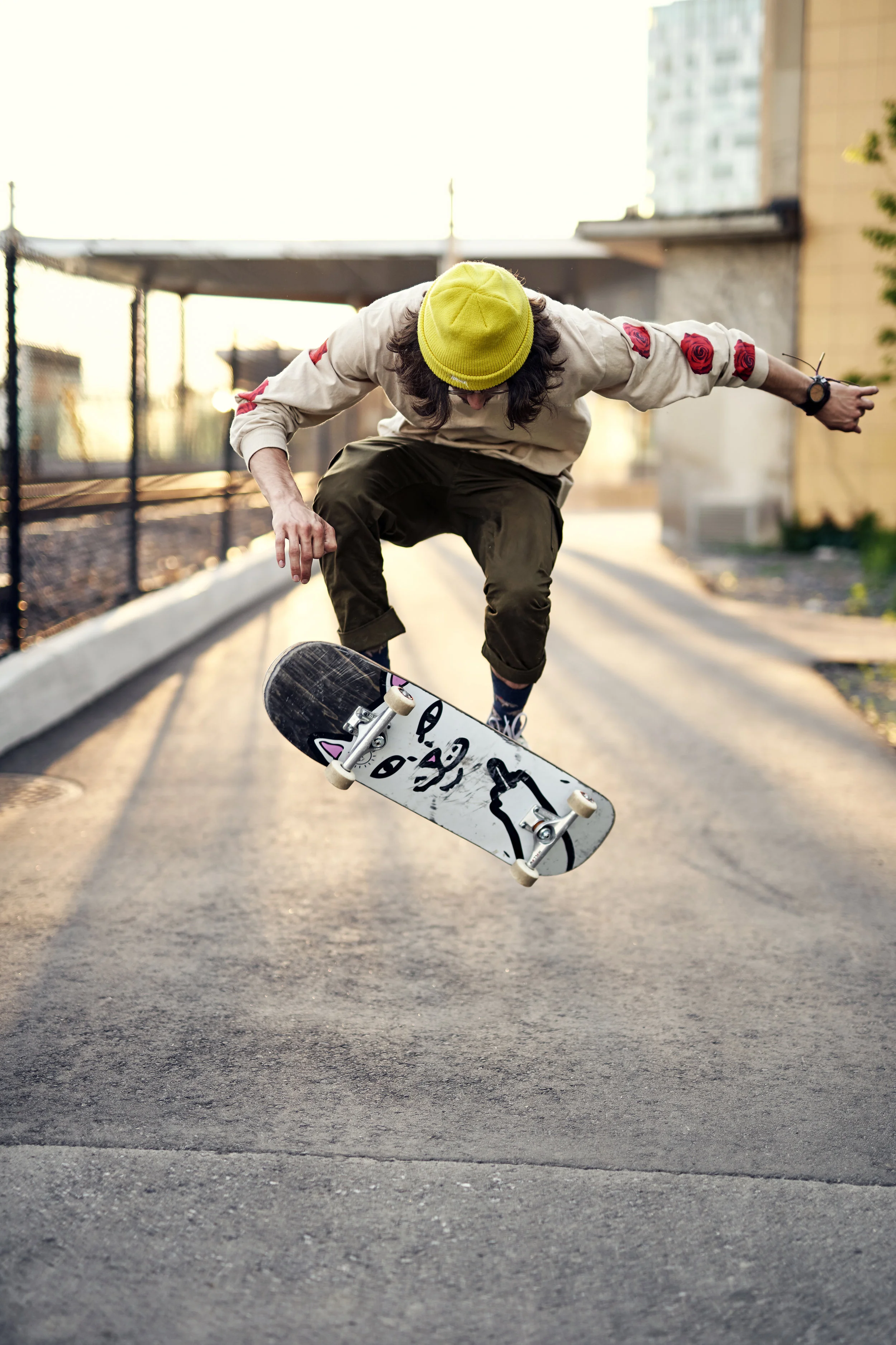 Skateboarder performing a trick on an outdoor paved path during sunset, wearing a yellow beanie, beige sweatshirt with red rose patches on sleeves, and dark pants.