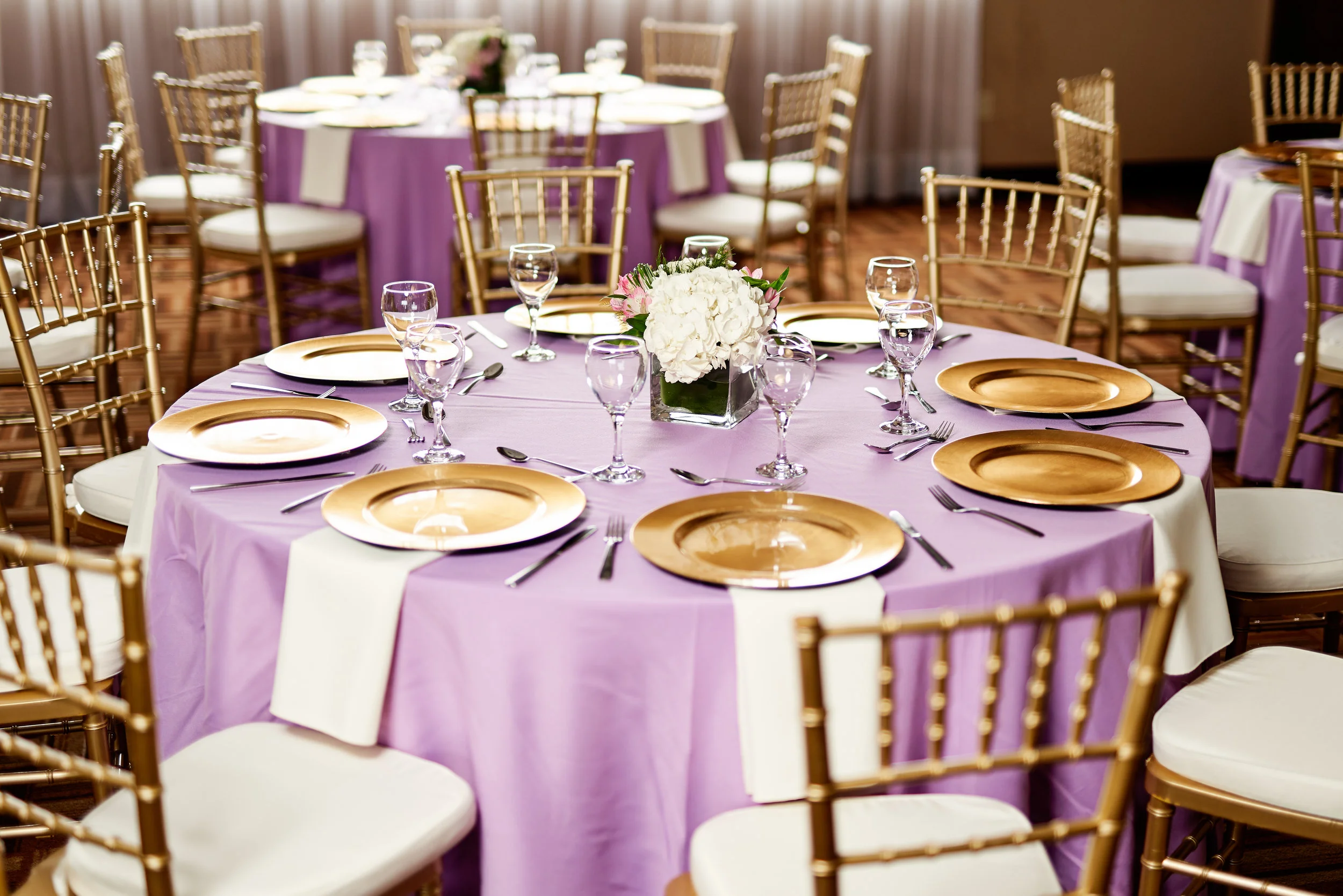 Round banquet table with purple tablecloth, gold plates, silverware, and water glasses, decorated with a white floral centerpiece, surrounded by gold chairs with white cushions in a banquet hall.