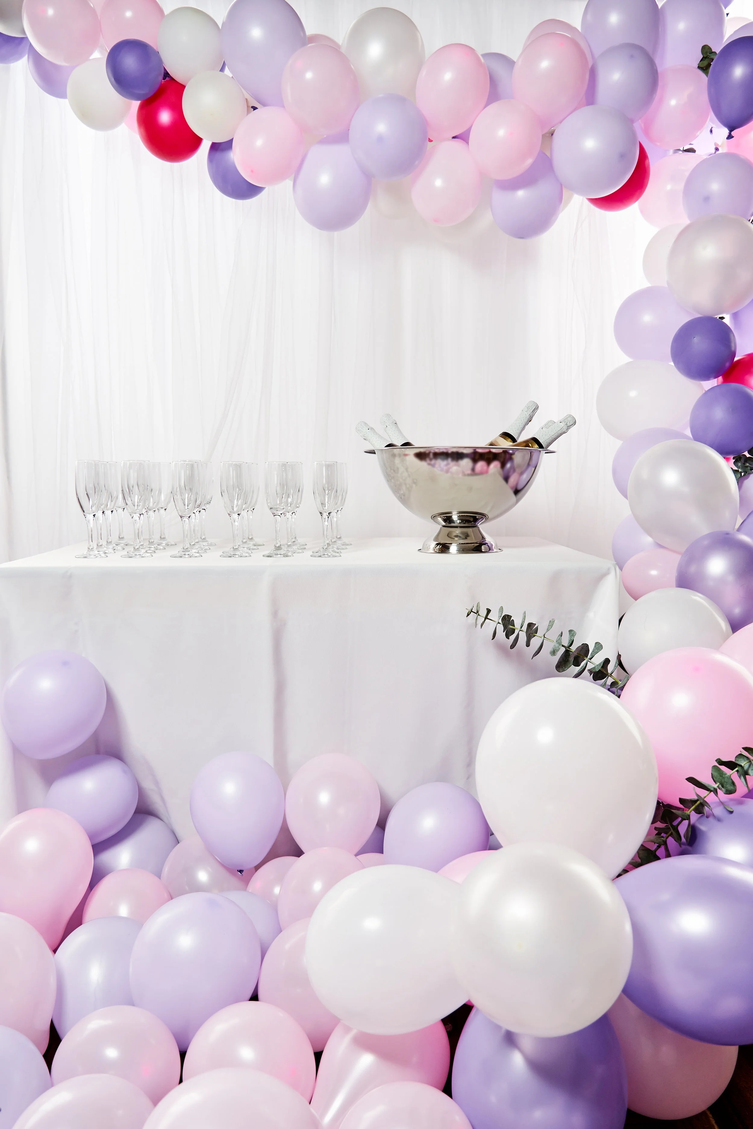 Decorative setup for a celebration with pastel-colored balloons in pink, lavender, and white surrounding a table with champagne glasses and a bowl of Champagne bottles, with a white curtain backdrop.