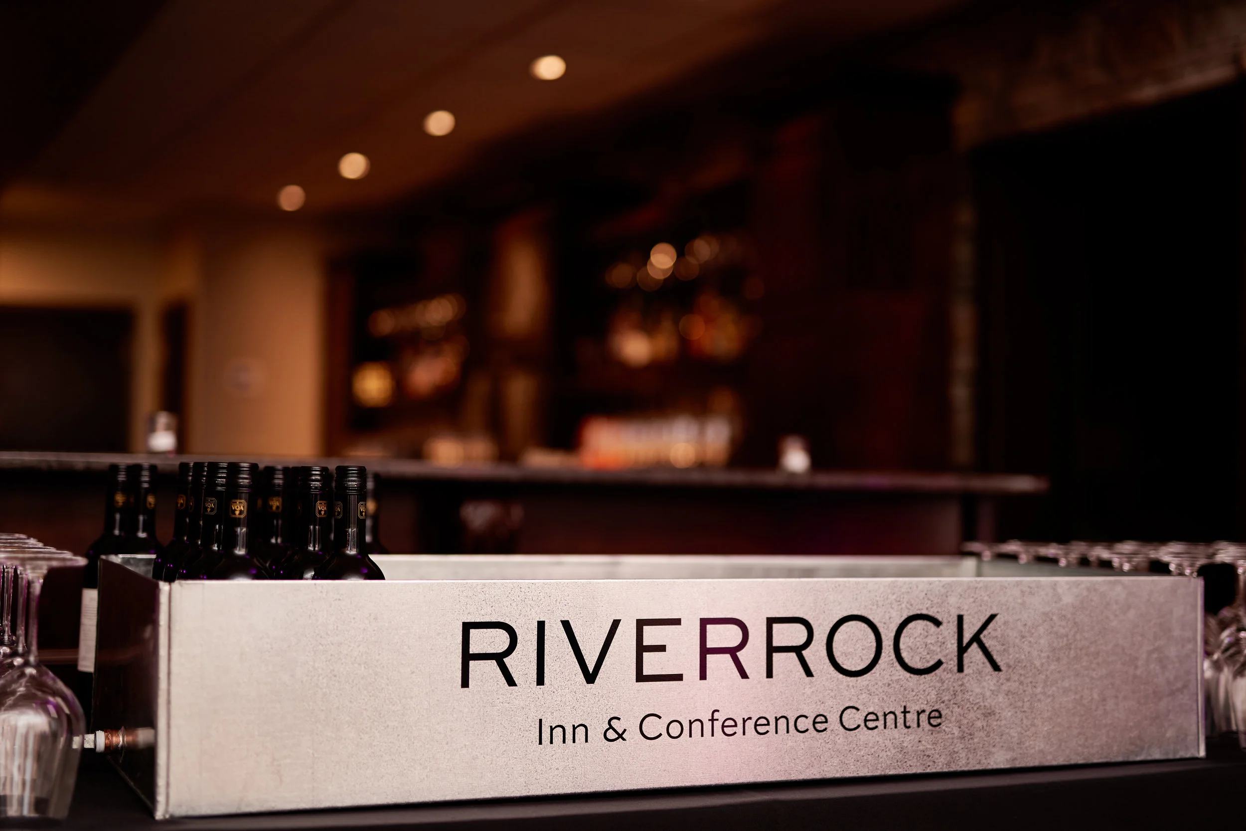 Bar with bottles of wine and glassware, sign reading 'RIVERROCK Inn & Conference Centre' in a dimly lit lounge area.
