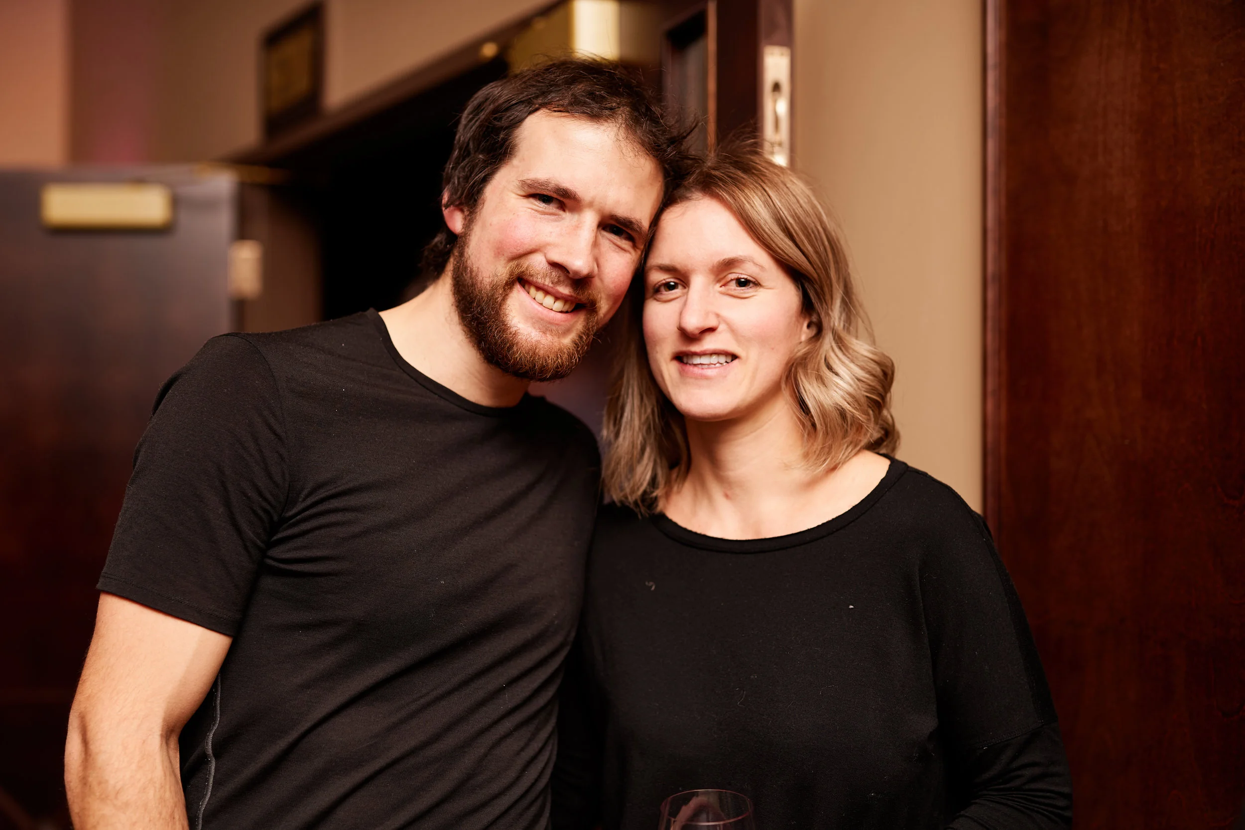 A smiling man and woman standing close together indoors, with the man slightly behind and to the left of the woman, both facing the camera.