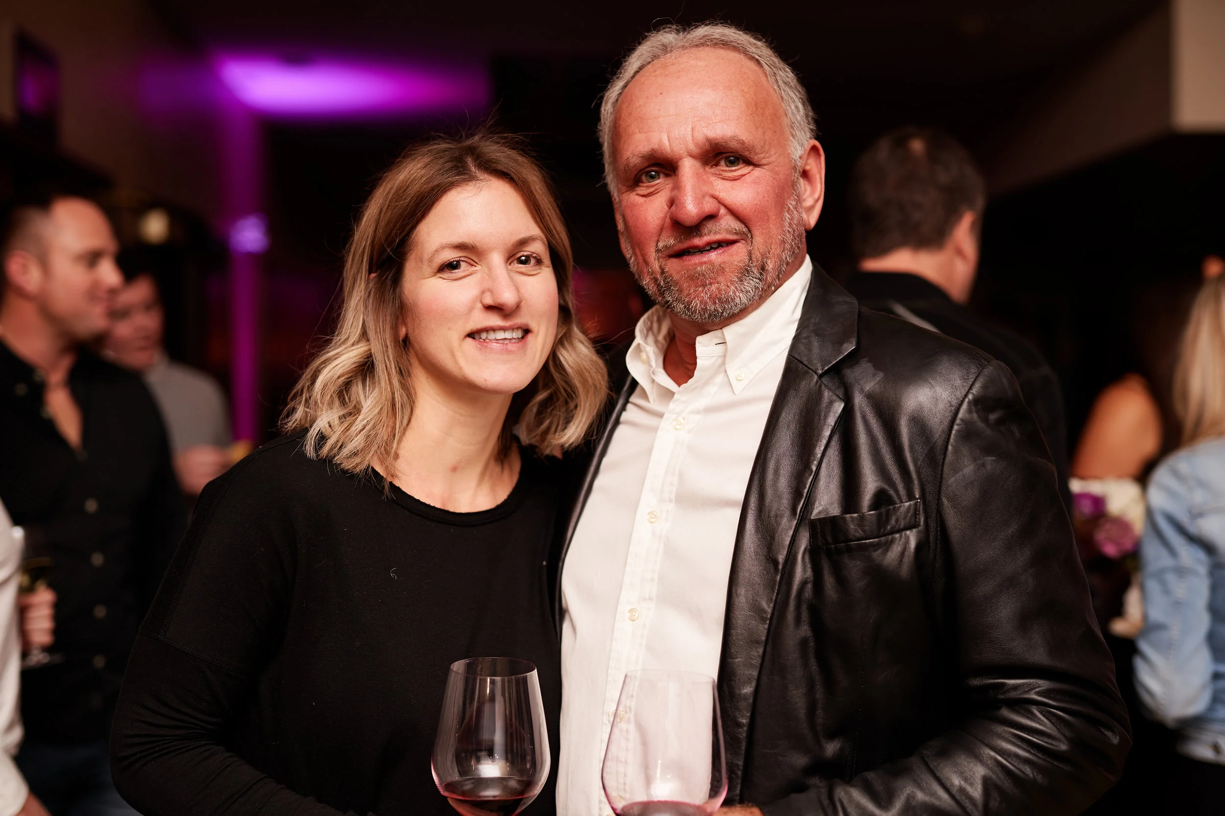 A woman and a man with gray hair and beard smiling while holding glasses of red wine at a social event.