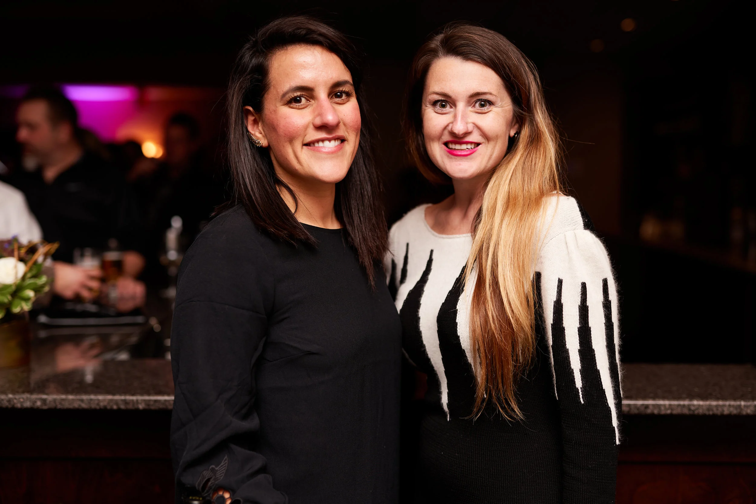 Two women smiling at a social event, standing close together in a dimly lit venue.