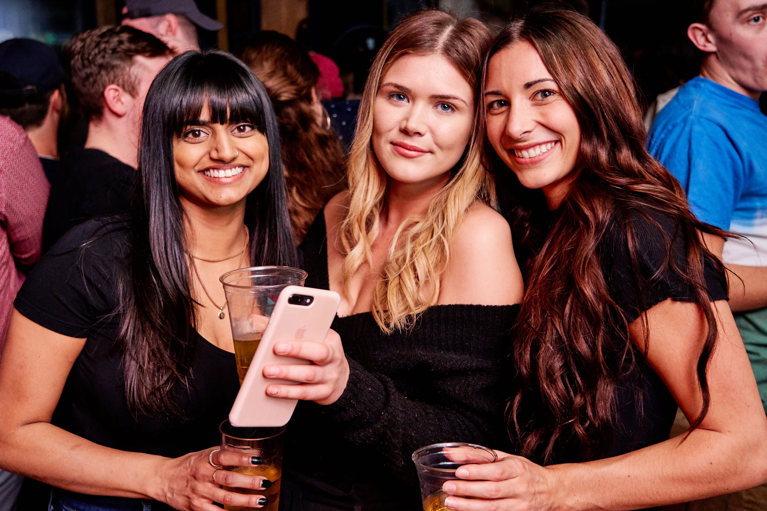 Three smiling women at a social event, holding drinks and taking a selfie together.