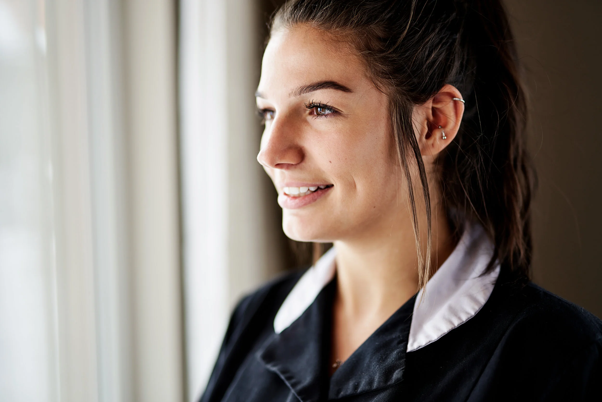A young woman with dark hair, wearing multiple earrings, smiling and looking out of a window in natural light.