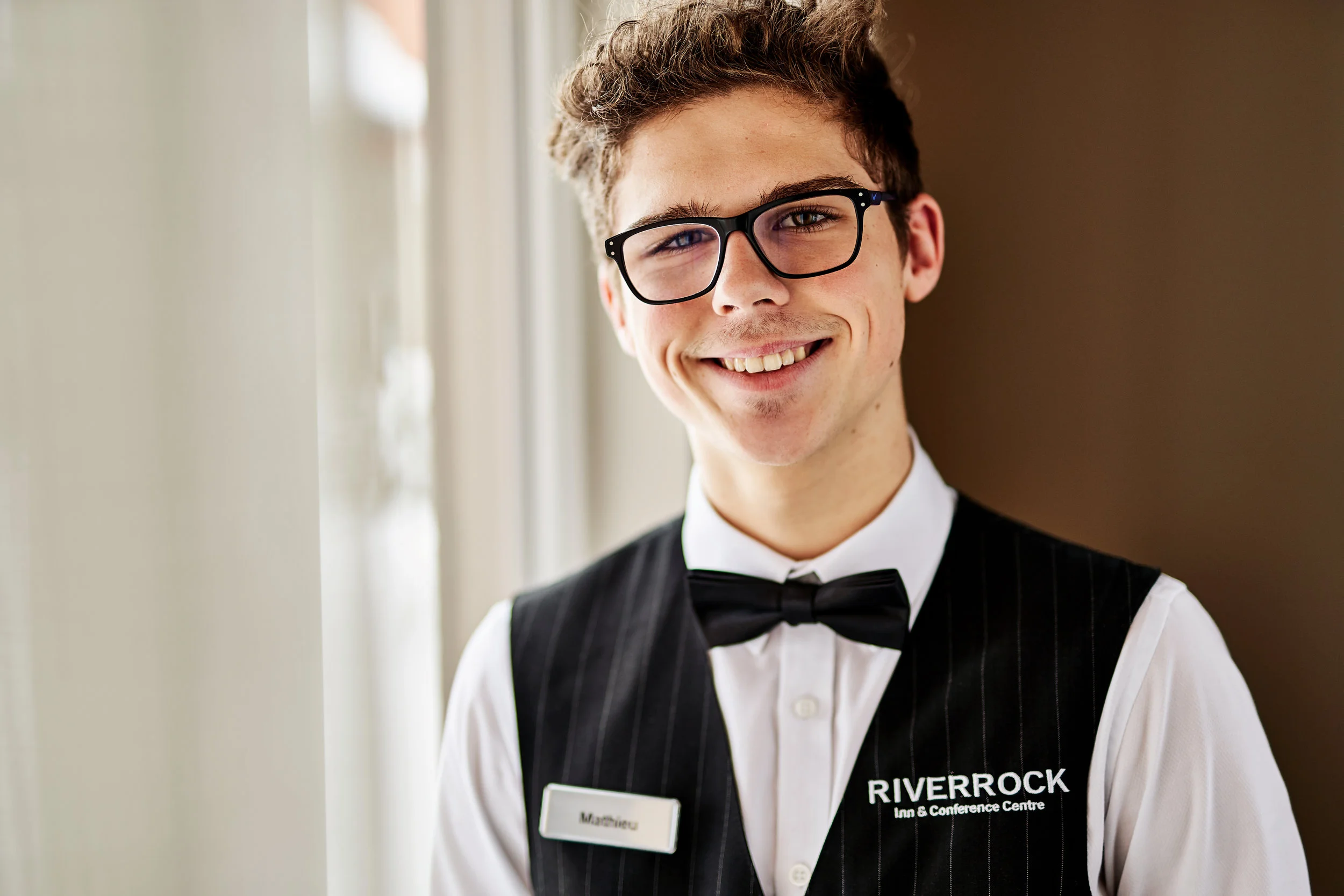 A young man with glasses, smiling, dressed in a tuxedo with a bow tie and a vest, standing near a window with sunlight, at the RiverRock Inn & Conference Centre.