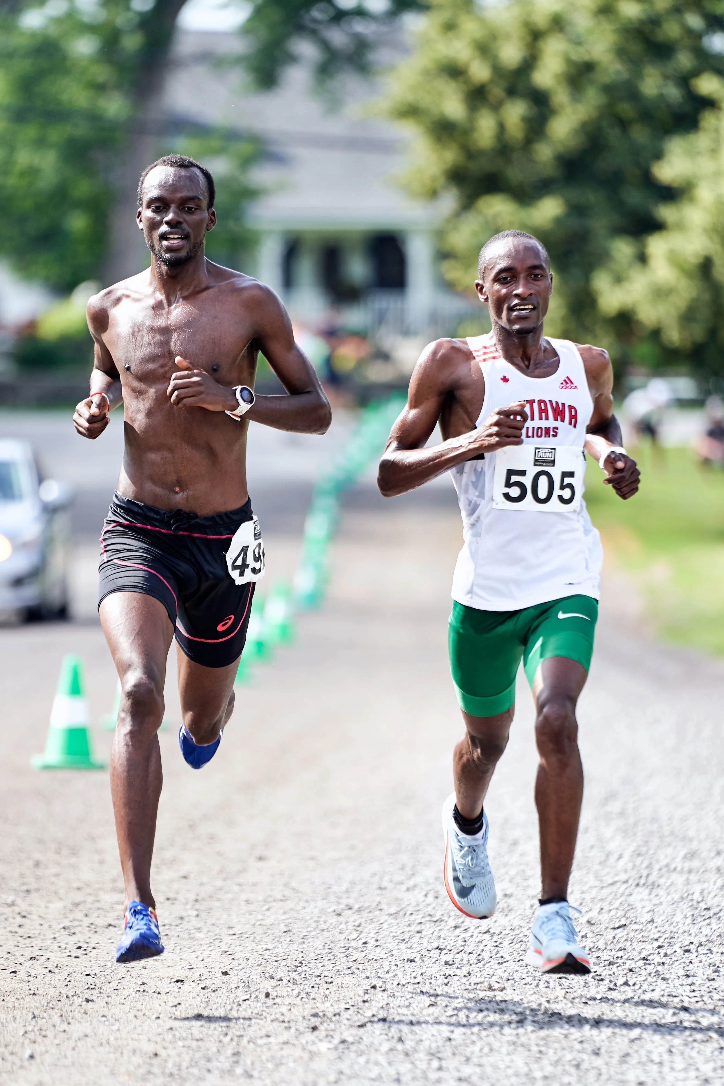 Two male runners are competing in a race, running on a gravel path with green cones along the side. One runner is shirtless, wearing black shorts with pink accents and a running watch, while the other wears a white tank top with red text and green sh