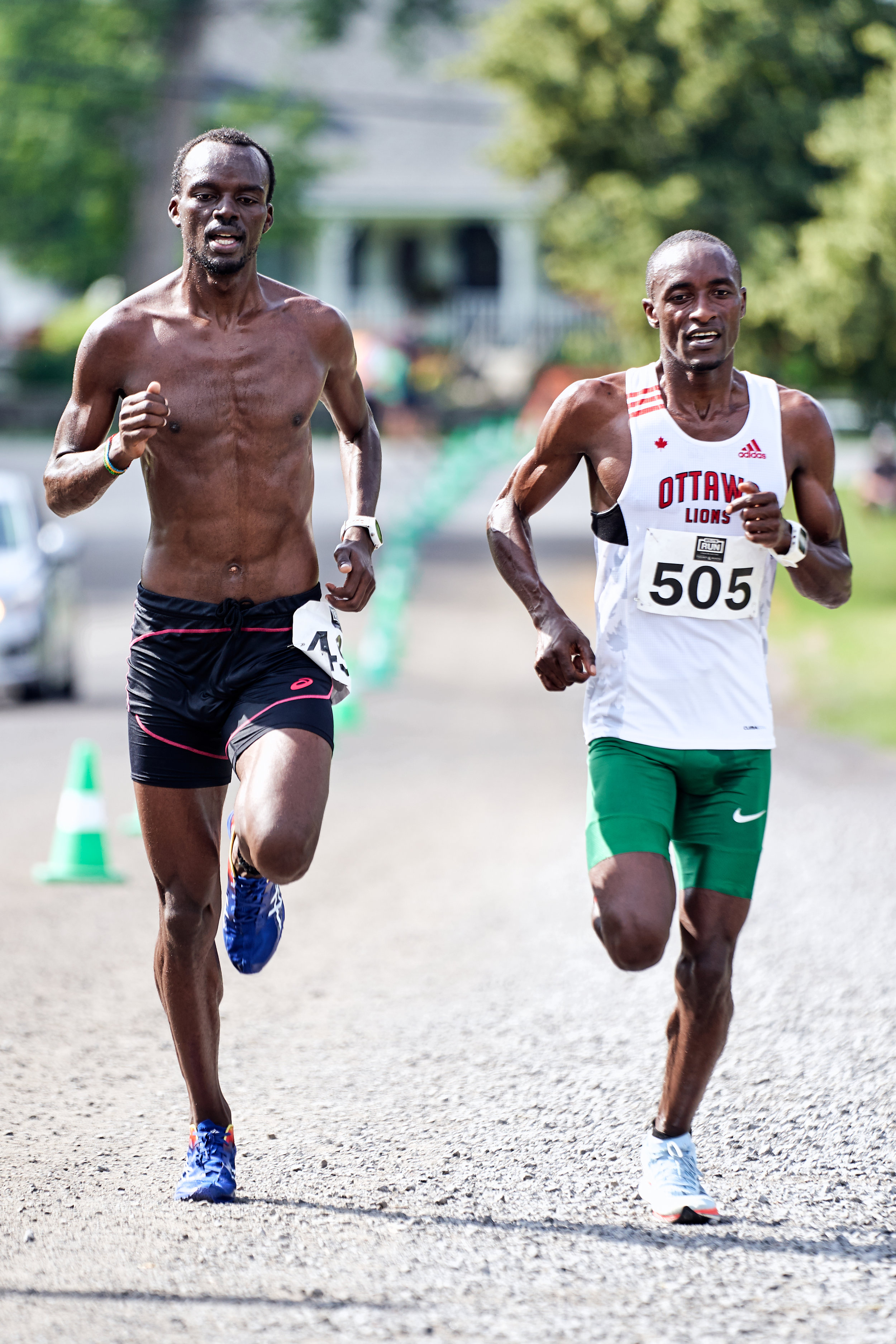 Two male runners competing in a race on a gravel path, with green trees and houses in the background. One runner is shirtless, wearing black shorts, and the other is wearing a white tank top and green shorts.
